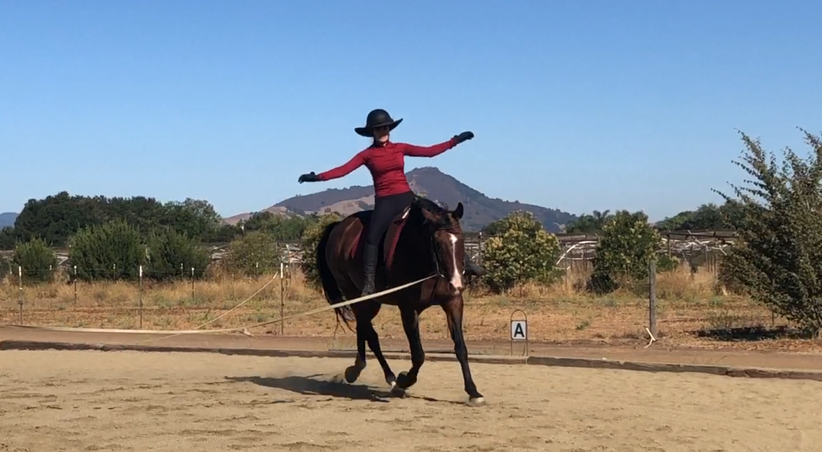 A woman riding a horse on a sandy outdoor riding arena, balancing with arms outstretched, wearing a wide-brimmed hat, red shirt, black pants, and gloves, with mountains and trees in the background under a clear blue sky.