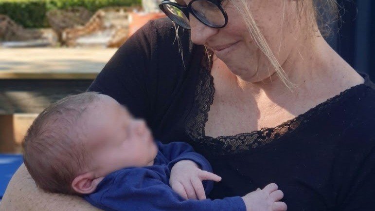 A woman holding a newborn baby outdoors, looking at the baby with affection.