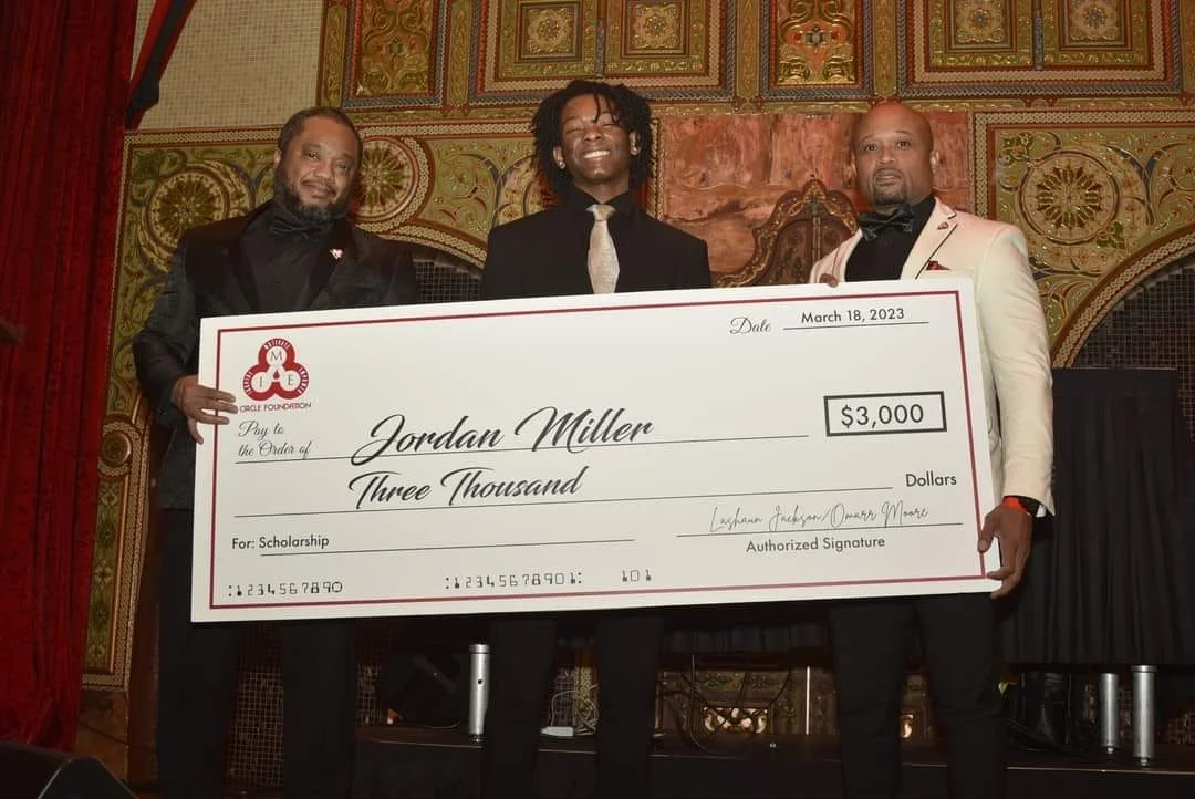Three men in formal attire holding a large ceremonial check for $3,000 made out to Jordan Miller, awarded as a scholarship, on a stage with ornate gold and red decor.