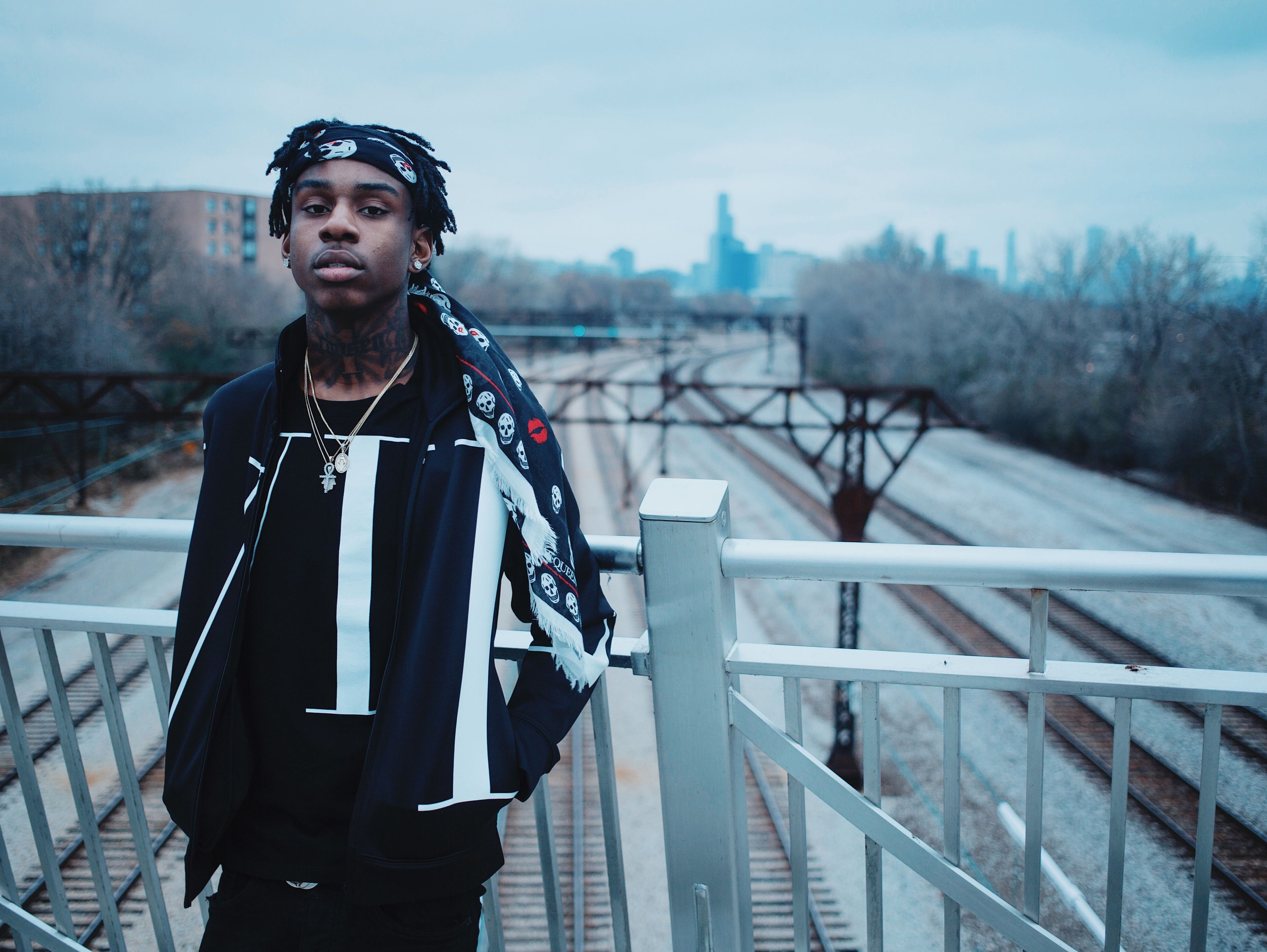 A young man with dreadlocks wearing a black jacket, a patterned scarf, and jewelry, standing on a bridge with city buildings and trees in the background during overcast weather.