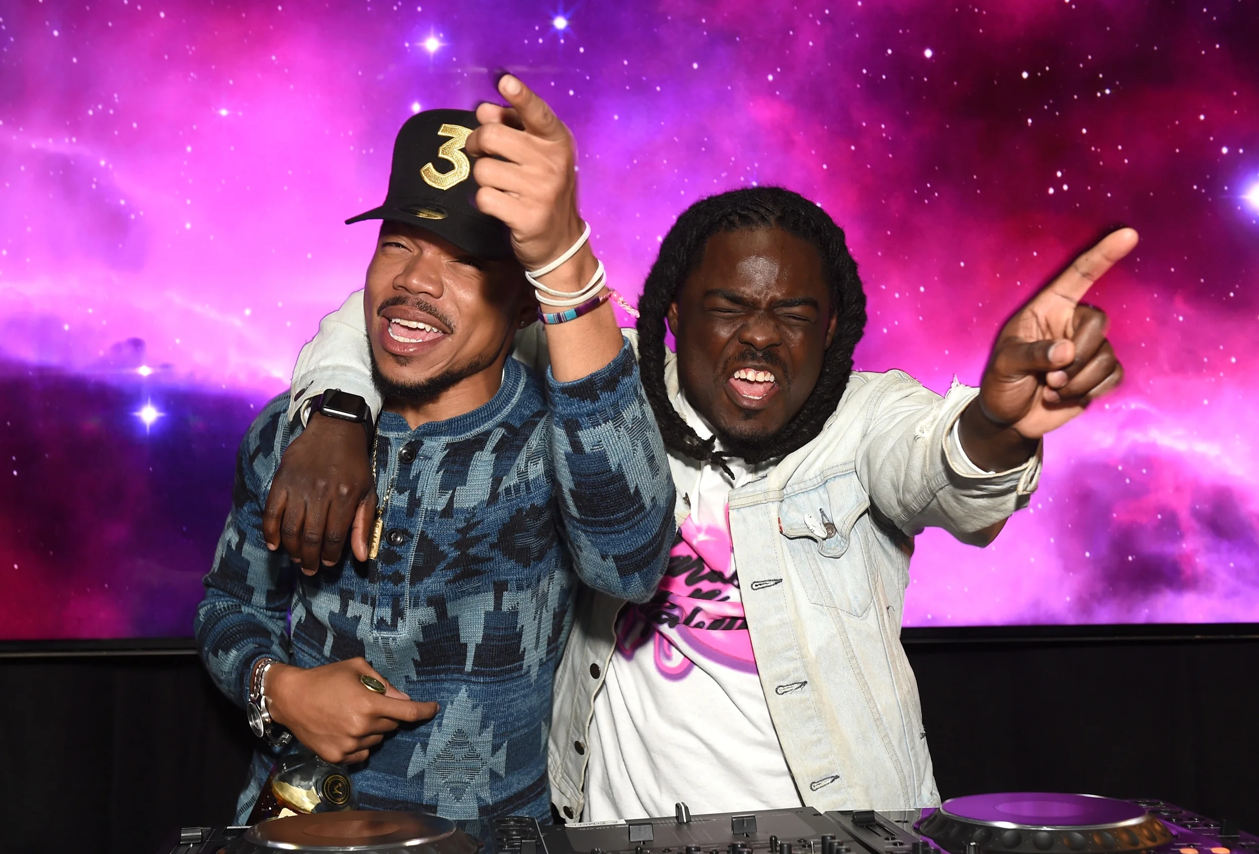 Two men are celebrating in front of a vibrant purple and pink galaxy backdrop. The man on the left is wearing a camouflage patterned jacket, holding a champagne glass, and adjusting a black cap with a gold number '3' on it. The man on the right has dreadlocks, is wearing a light-colored jacket over a pink and black shirt, and is pointing with an energetic expression. There is DJ equipment in front of them.