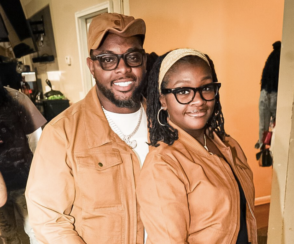 A smiling man and woman both dressed in matching tan jackets at an indoor gathering.