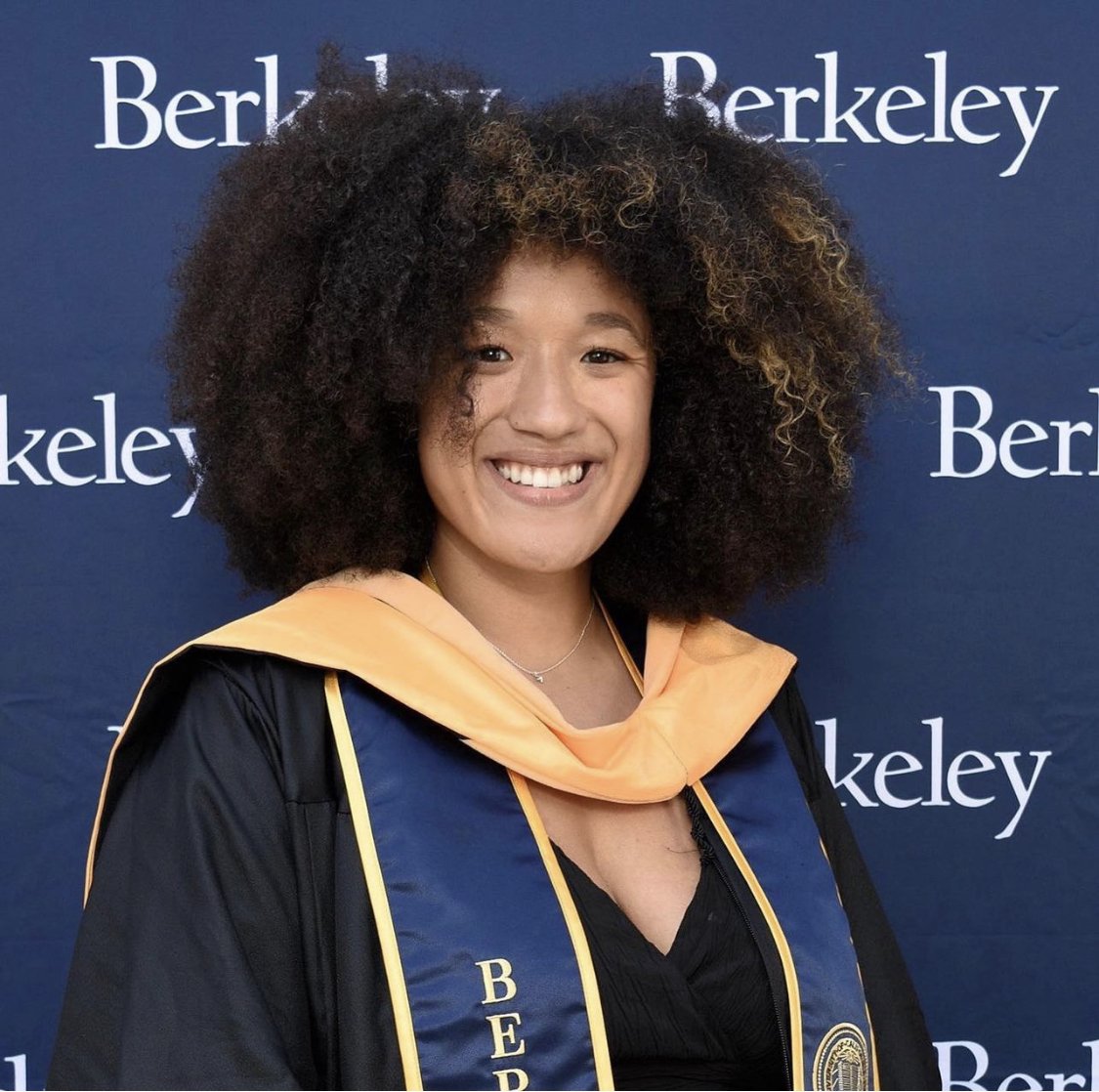 A woman smiling in graduation attire standing in front of a blue backdrop with white text that reads 'Berkeley' repeatedly.