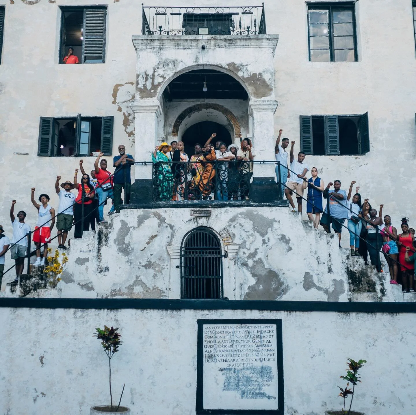 A group of diverse people standing on a historic, weathered white building with open windows and an arched doorway, raising their fists in a gesture of solidarity or protest.