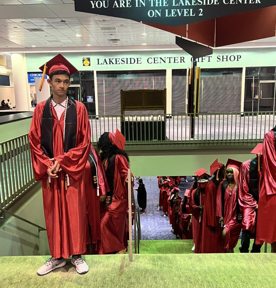 Group of graduates in red caps and gowns on a staircase inside a shopping mall, with a young man at the top looking to the side. The background shows a sign for Lakeside Center Gift Shop and other mall storefronts.