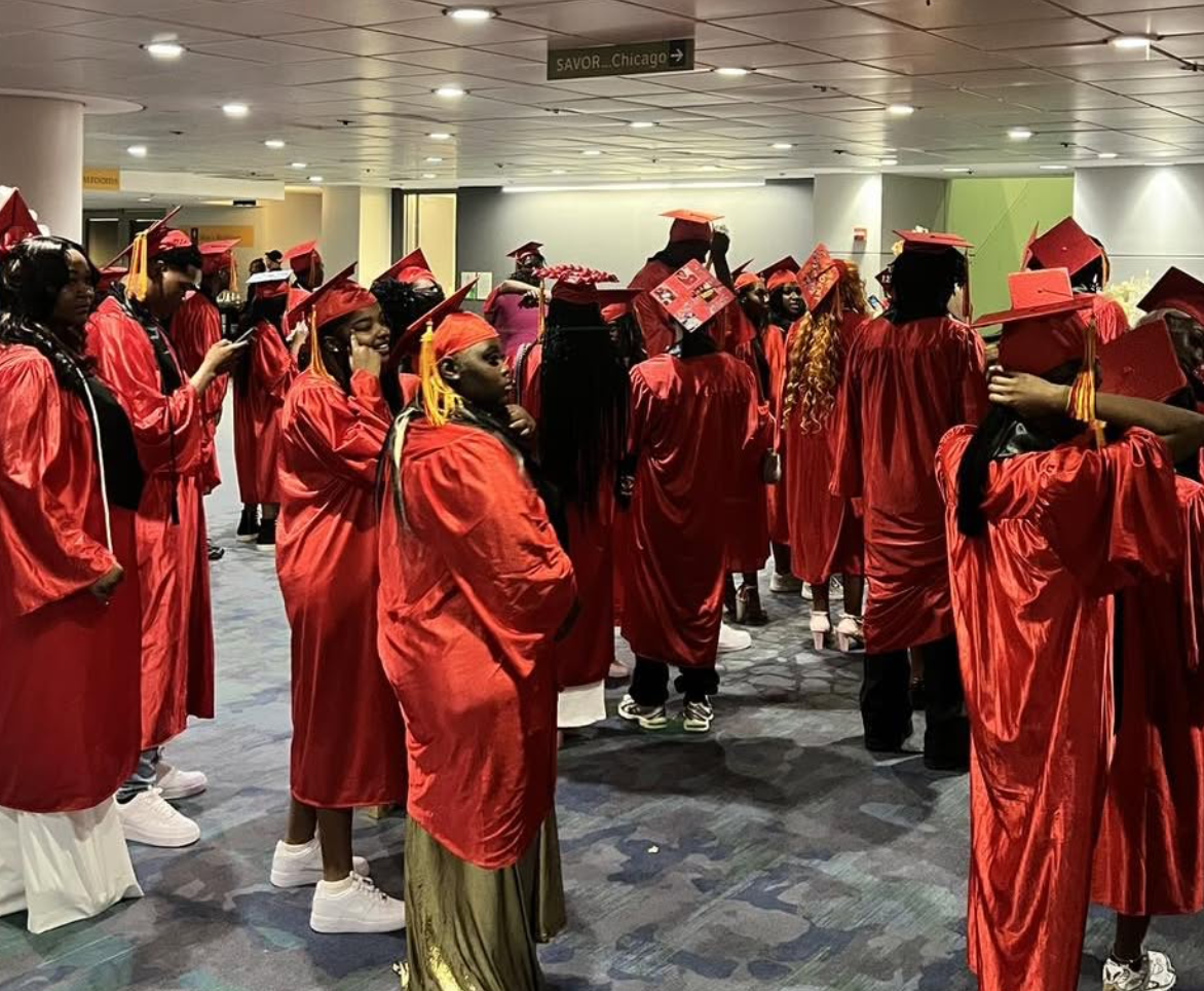 Group of graduates in red caps and gowns waiting indoors, some adjusting hats or chatting, at a graduation ceremony.