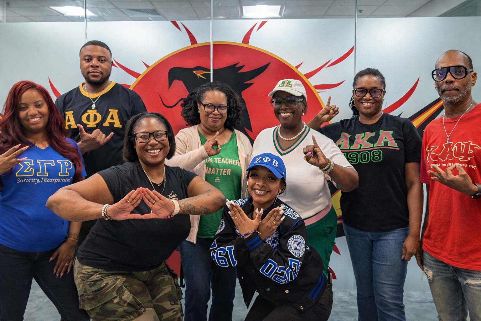 Group of diverse women and men posing with hand signs in front of a red and black eagle logo, some wearing sorority and fraternity shirts.