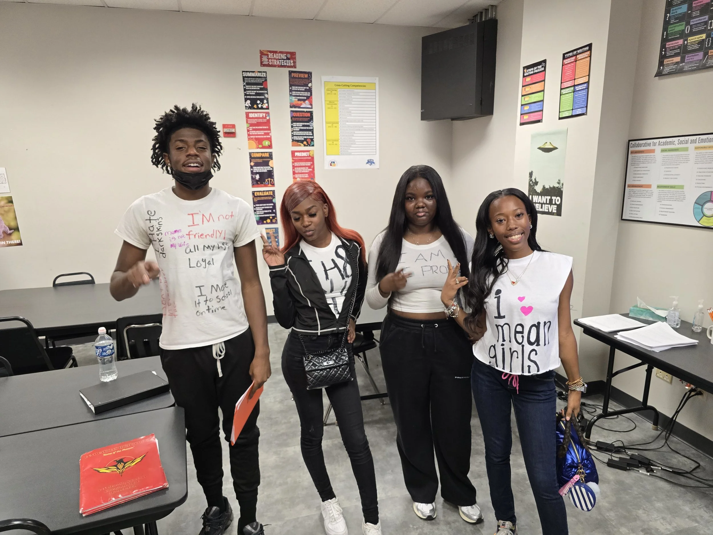 Four young women standing together in a classroom, smiling and making peace signs. They are surrounded by classroom posters and tables with notebooks and water bottles.