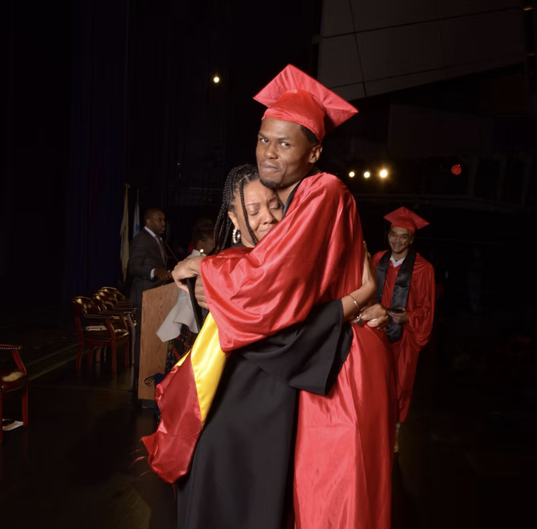 A young man in a red graduation gown and cap hugging a woman who appears to be emotional, during a graduation ceremony. Another graduate in a similar gown is visible in the background.