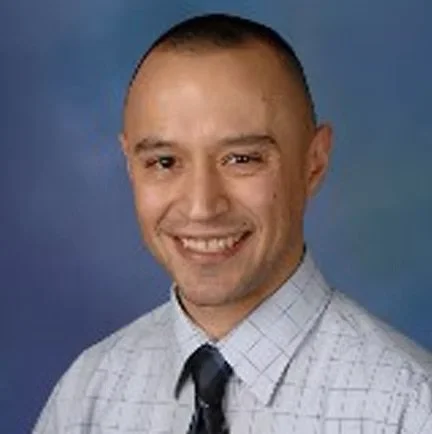 A man with short hair and a friendly smile, wearing a light-colored, checkered dress shirt and a dark tie against a blue background.