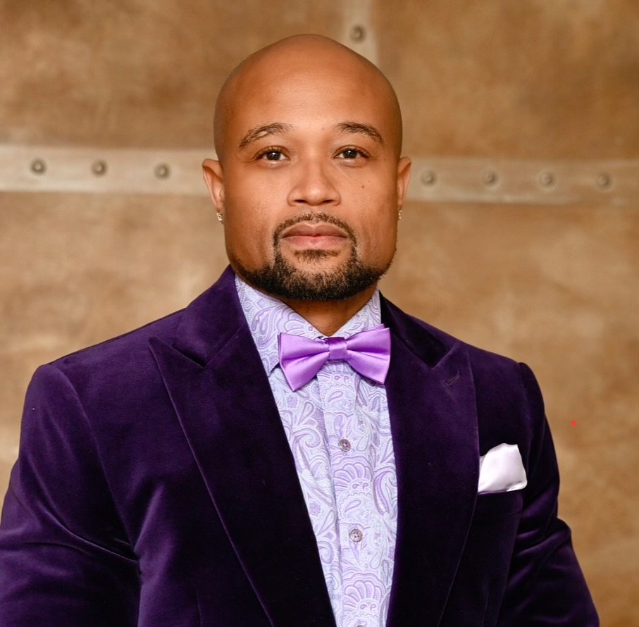 A man with a shaved head and goatee, wearing a purple velvet blazer, patterned shirt, matching purple bow tie, and small earrings, standing in front of a wooden background.
