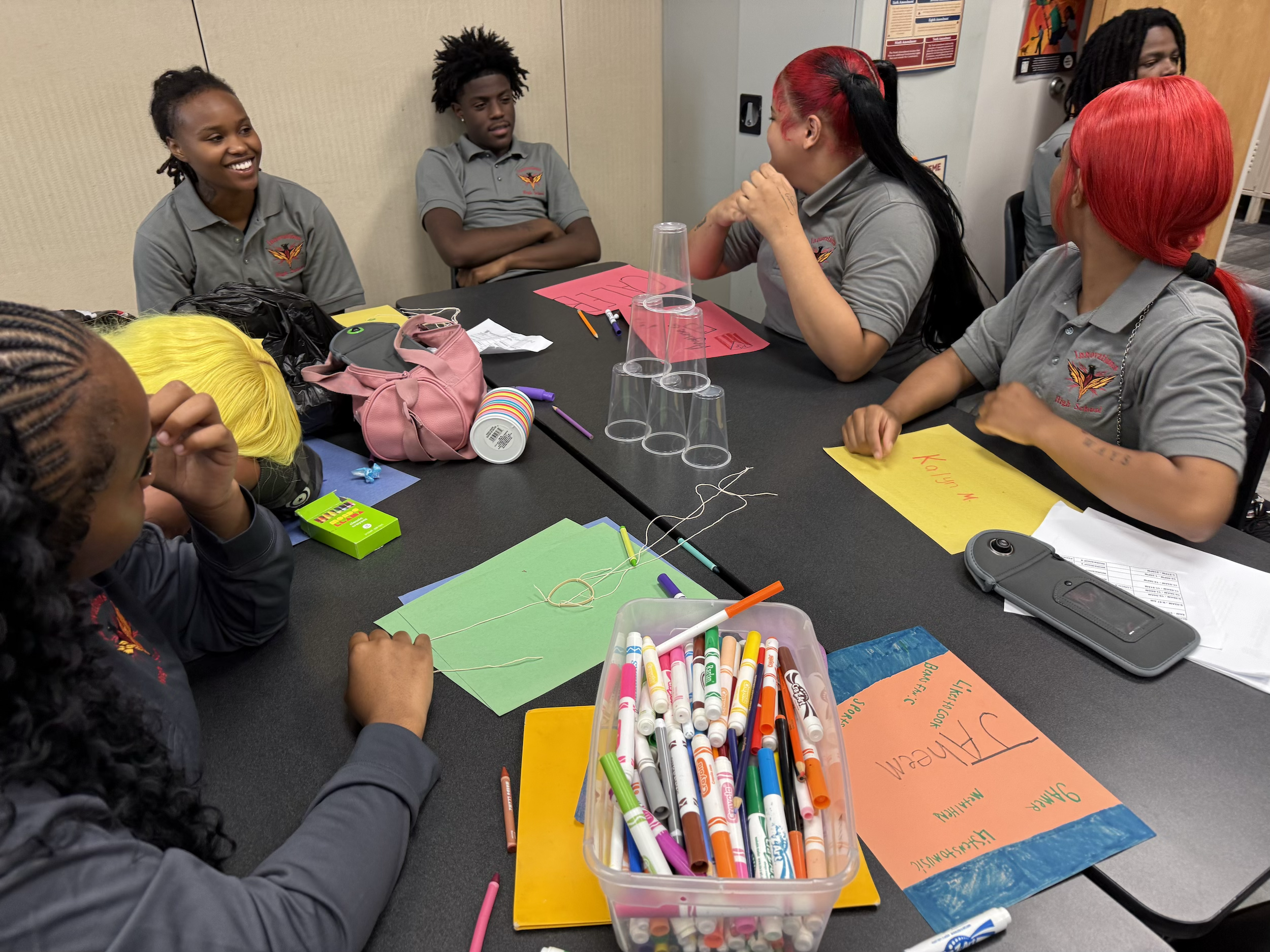 A group of young people sitting at a table, engaging in a discussion. They are wearing gray shirts with a logo, and there are colorful posters, markers, and cups on the table.