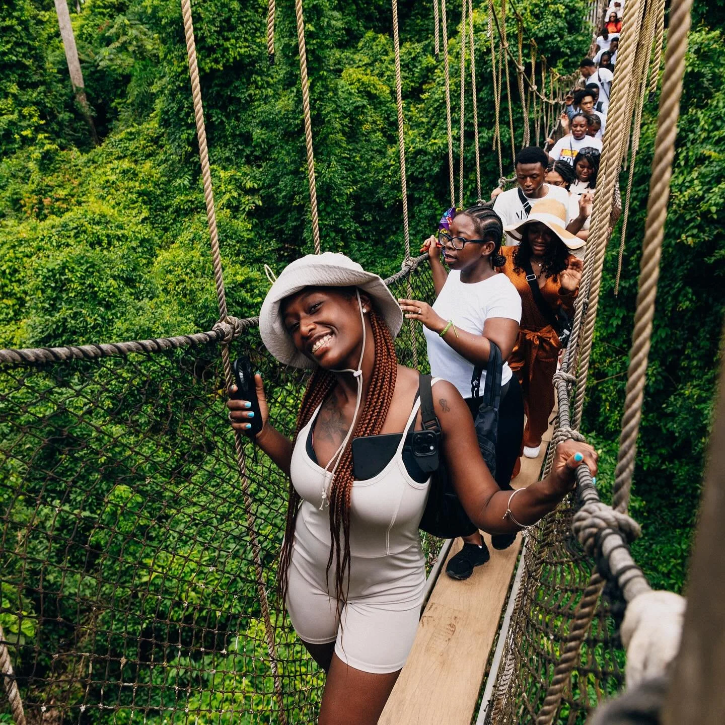People walking on a rope bridge through a lush green forest, smiling and holding onto the side ropes.