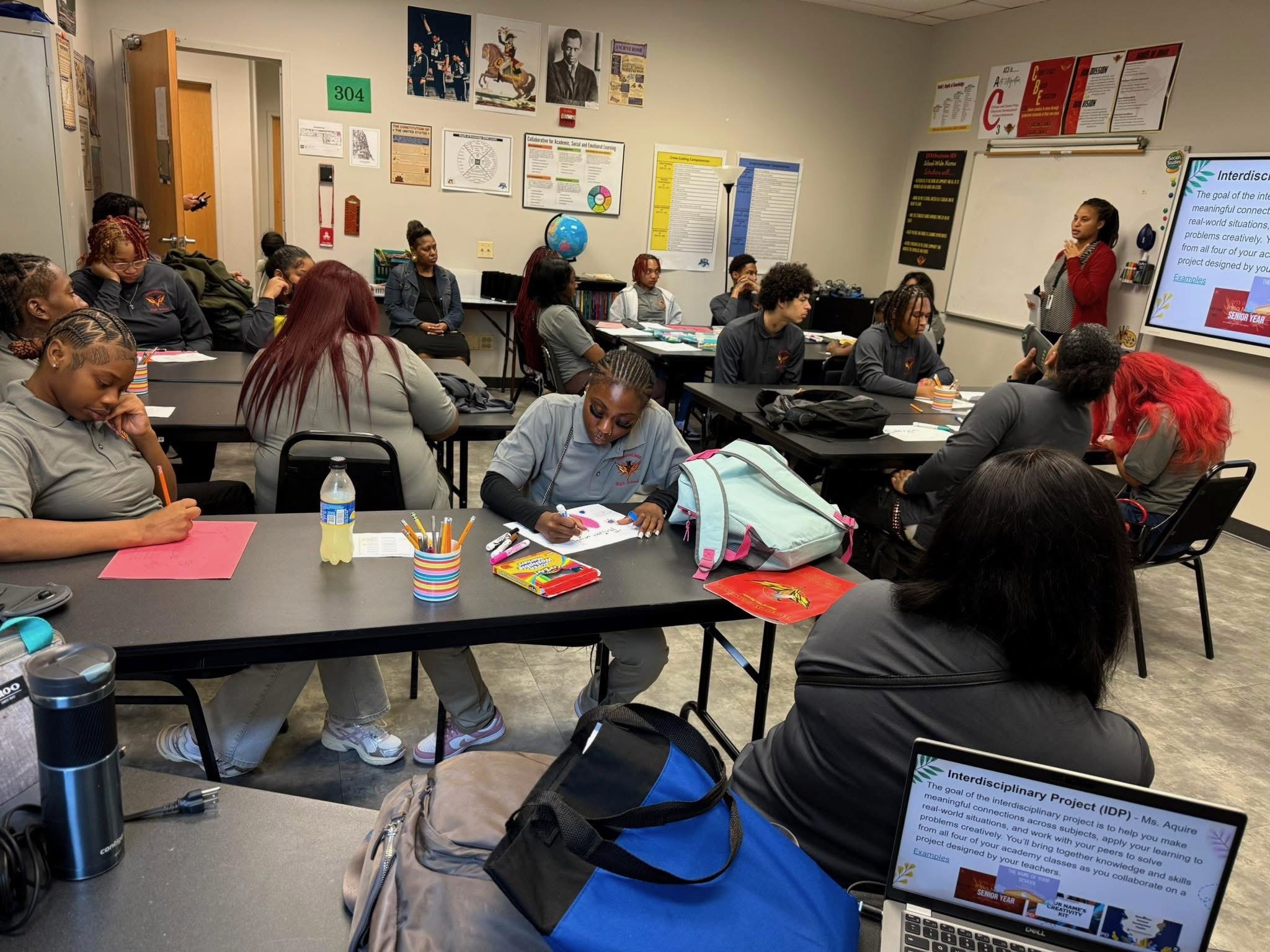 A classroom filled with students sitting at tables, some taking notes and others listening attentively to a teacher at the front. The teacher is pointing at a large screen displaying a presentation about interdisciplinary projects. The room has educa