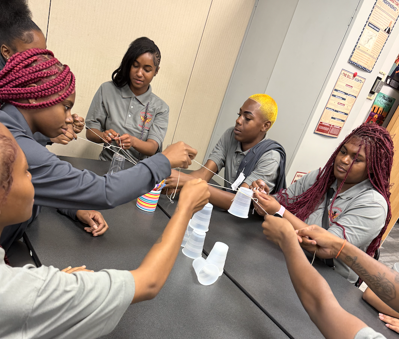 Group of young women participating in a hands-on STEM activity at a table, working with cups and strings.
