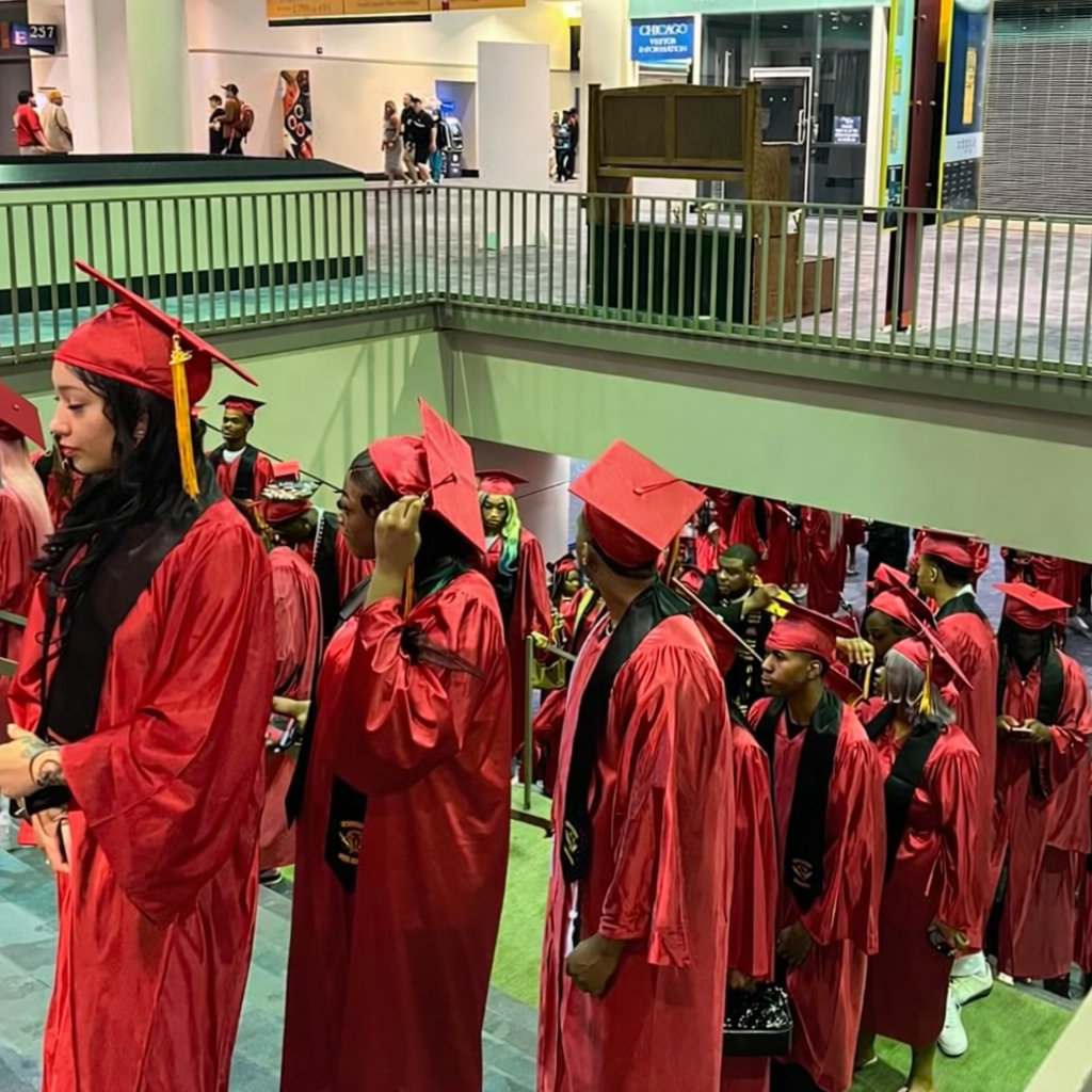 Group of students in red graduation gowns and caps standing in line inside a building, likely at a graduation ceremony.