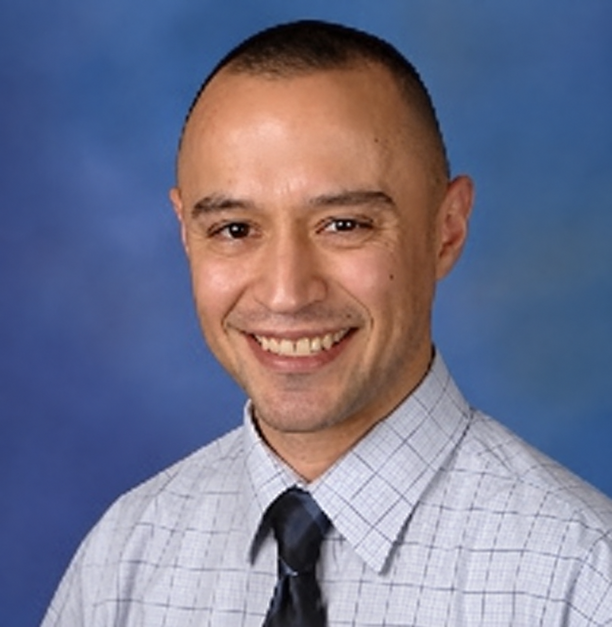 A man with short hair and a friendly smile, wearing a light-colored, checkered dress shirt and a dark tie against a blue background.