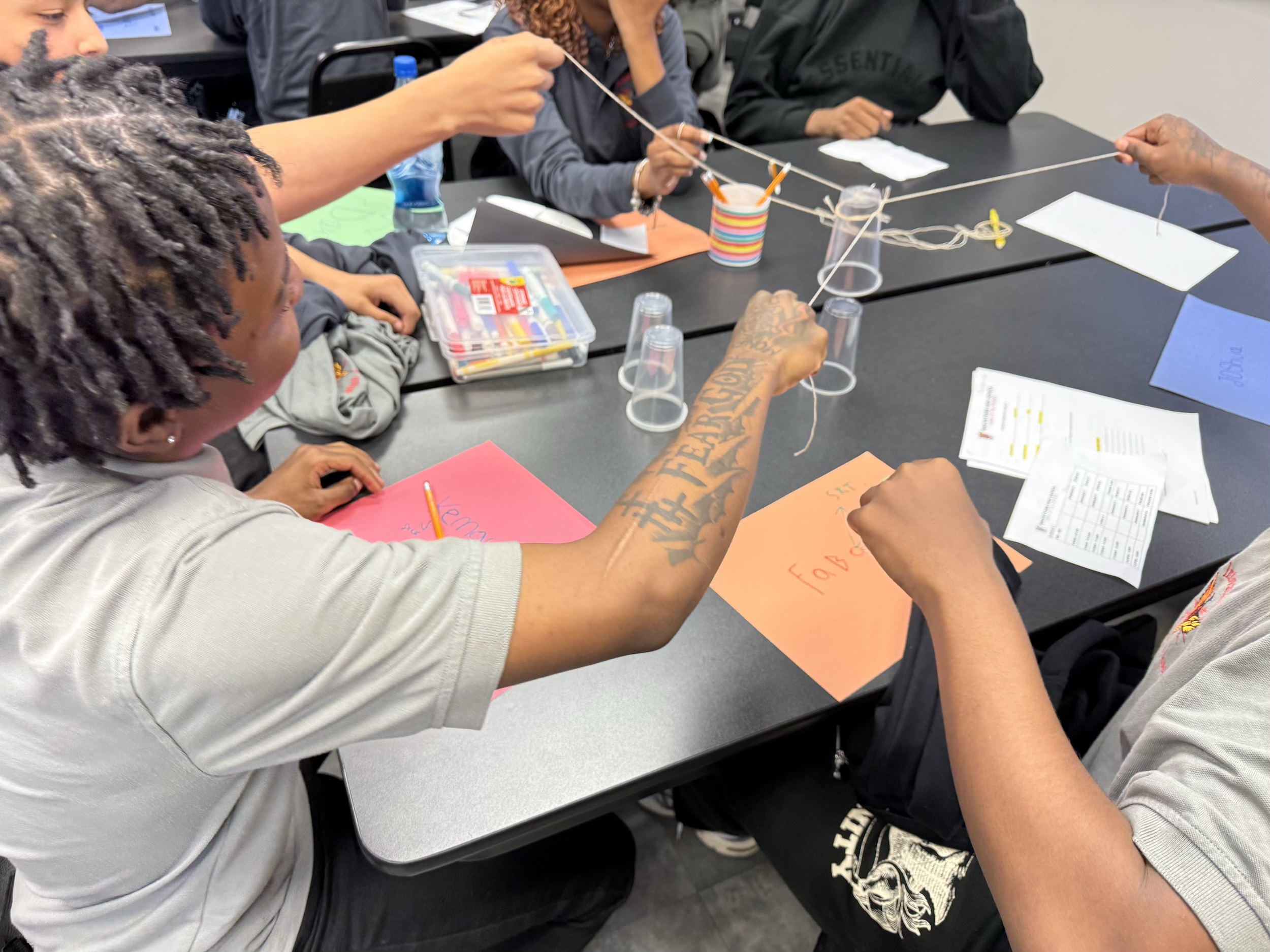 Several people sitting at a table participating in a team-building activity involving string and plastic cups, with various papers and supplies on the table.