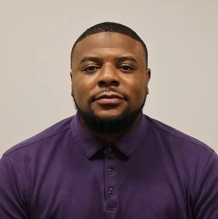 A man with short hair and a beard wearing a purple collared shirt, looking serious, plain gray background.