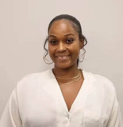 Portrait of a smiling Black woman with hoop earrings, wearing a white blouse and a gold necklace, against a plain gray background.