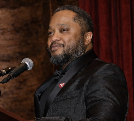 Man in black tuxedo speaking at a microphone on a stage with a red curtain background.