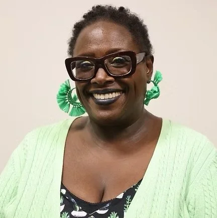 A woman with short curly hair, dark glasses, and large green earrings, smiling and wearing a light green cardigan over a patterned black and white top.