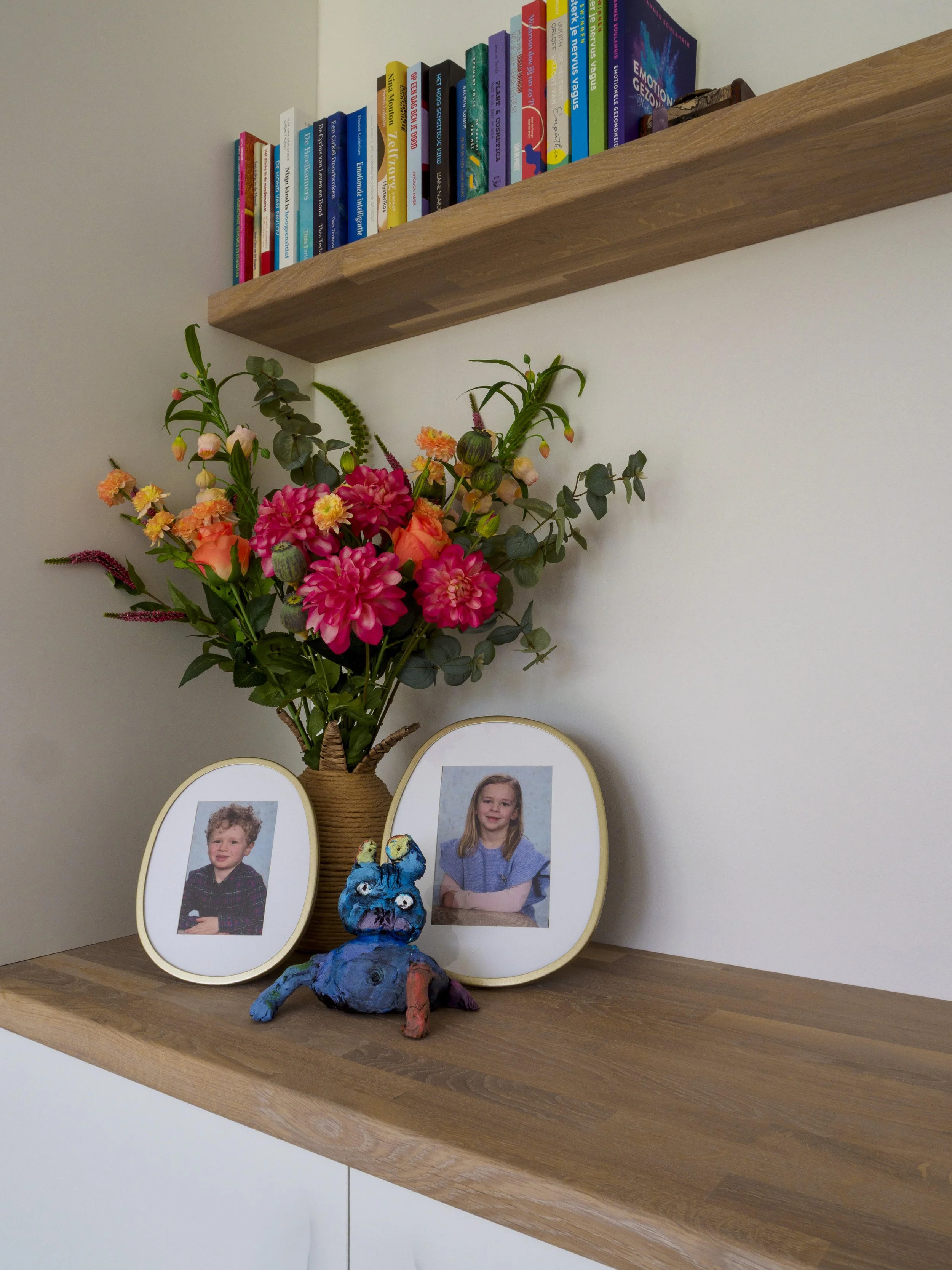 Een houten plank met daarop een bloemenvaas met kleurrijke bloemen, twee fotoframes met foto's van kinderen, en een kleurrijk knuffeldier dat op de plank staat.