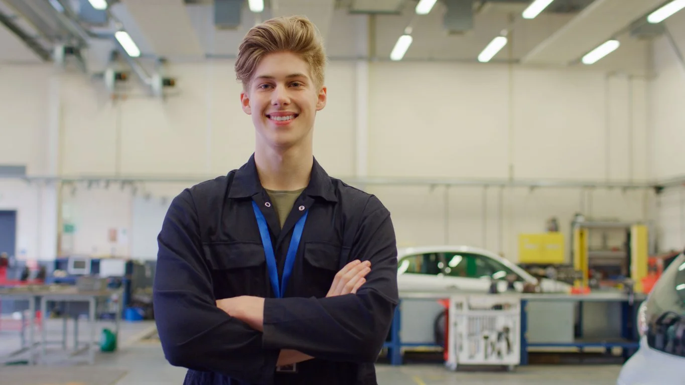 A smiling young person with blond hair in dark workwear stands confidently with folded arms in a bright automotive workshop, with cars and equipment behind him, reflecting occupational therapy support for meaningful employment.