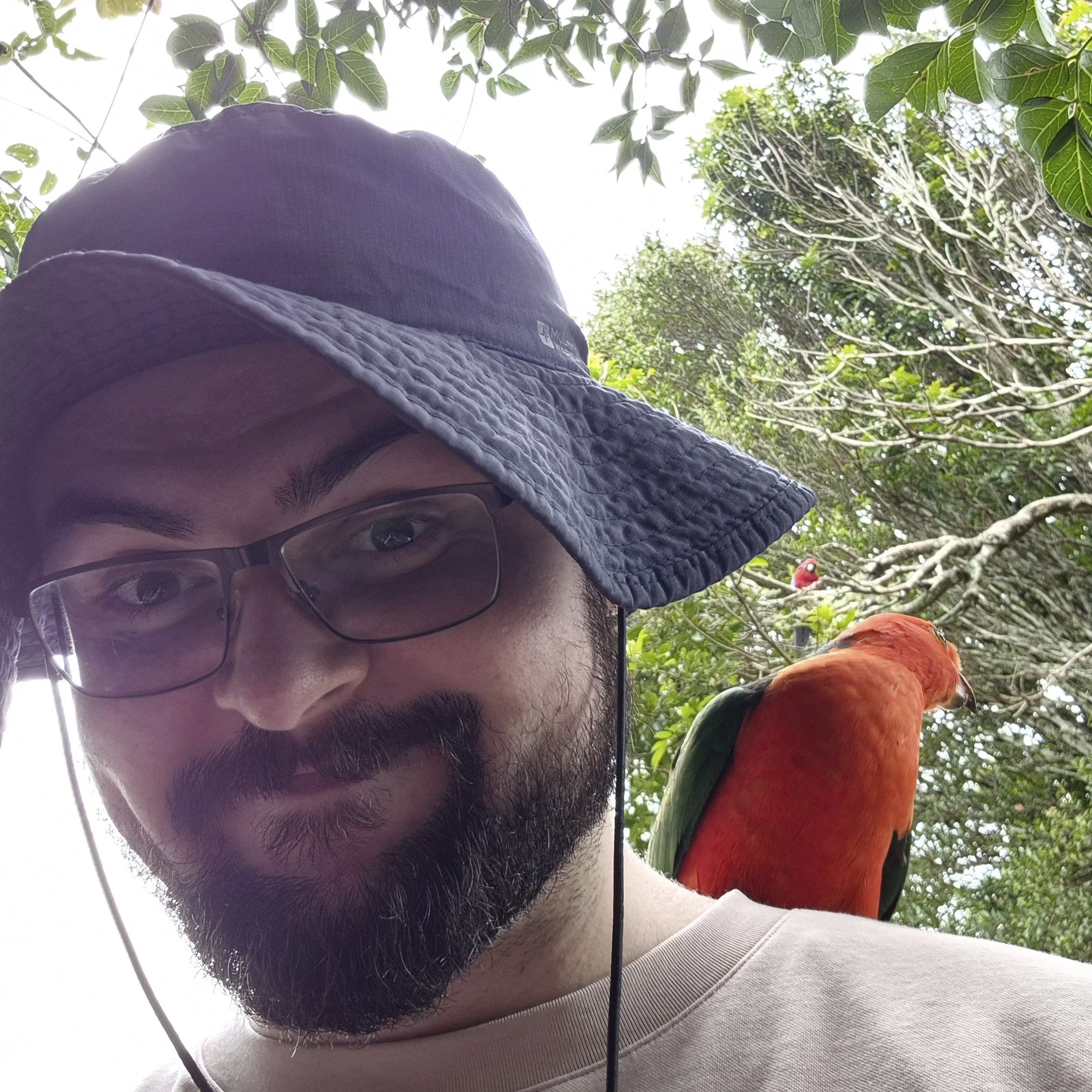 Outdoor photo of Callum, Education and Support Coach, smiling in glasses and a sunhat with a colourful parrot on his shoulder, reflecting his friendly, approachable and engaging support style.