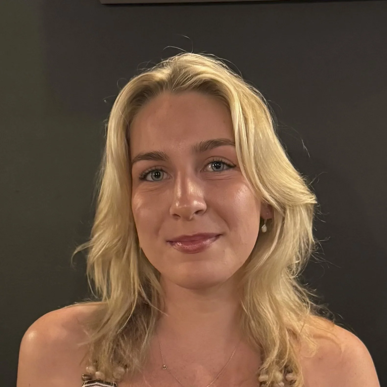 Professional headshot of Olivia, Employment and Daily Living Coach, smiling with blonde hair against a dark background, representing approachable, person-centred support for employment, everyday living skills, confidence and independence.