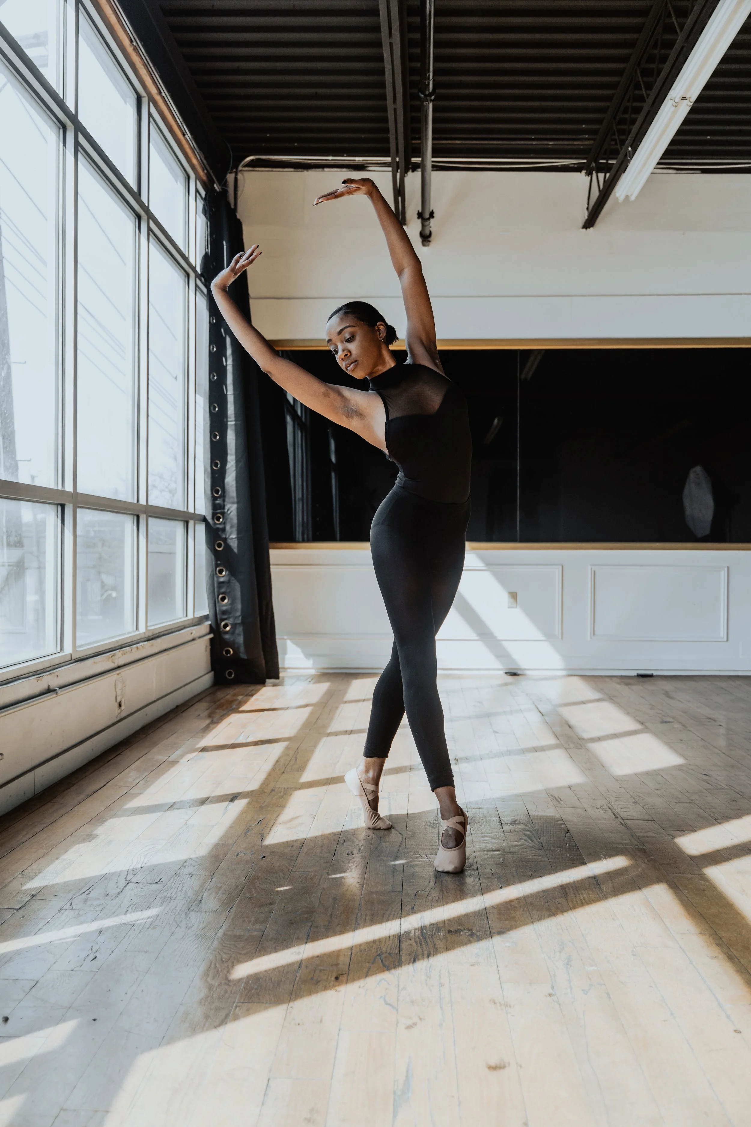 A female ballet dancer practicing in a dance studio with large windows and wooden floors.