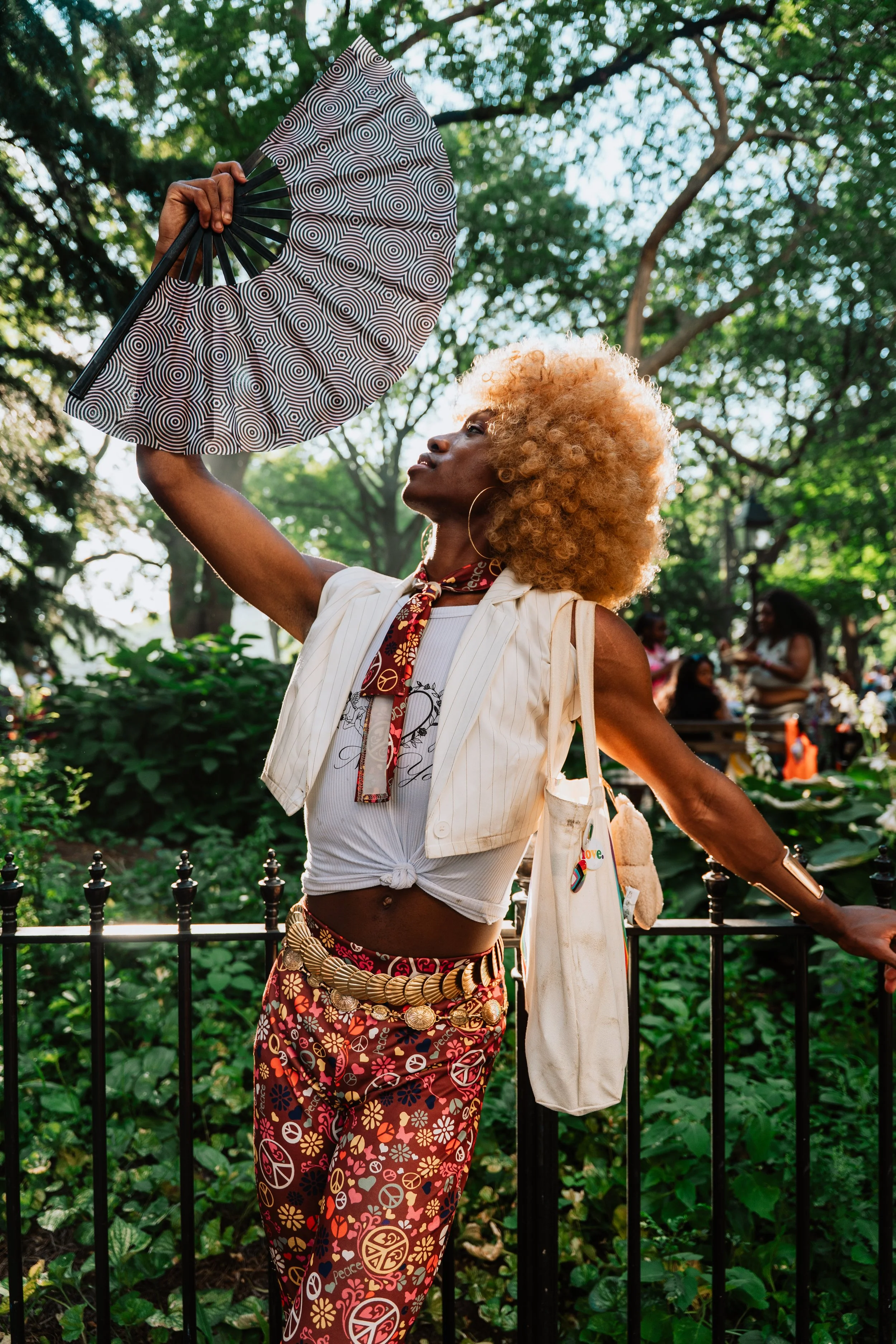 A person with blonde curly hair holds a patterned hand fan above their head in a park with green trees in the background.