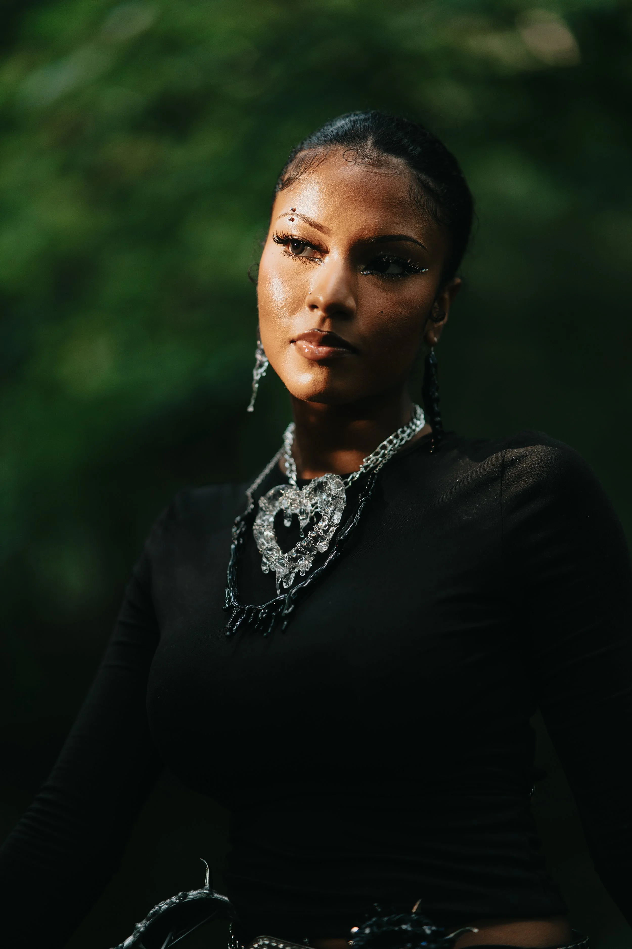 A woman with dark hair styled back, wearing elaborate silver jewelry including necklaces, earrings, and accessories, dressed in black, and standing outdoors with blurred green foliage in the background.