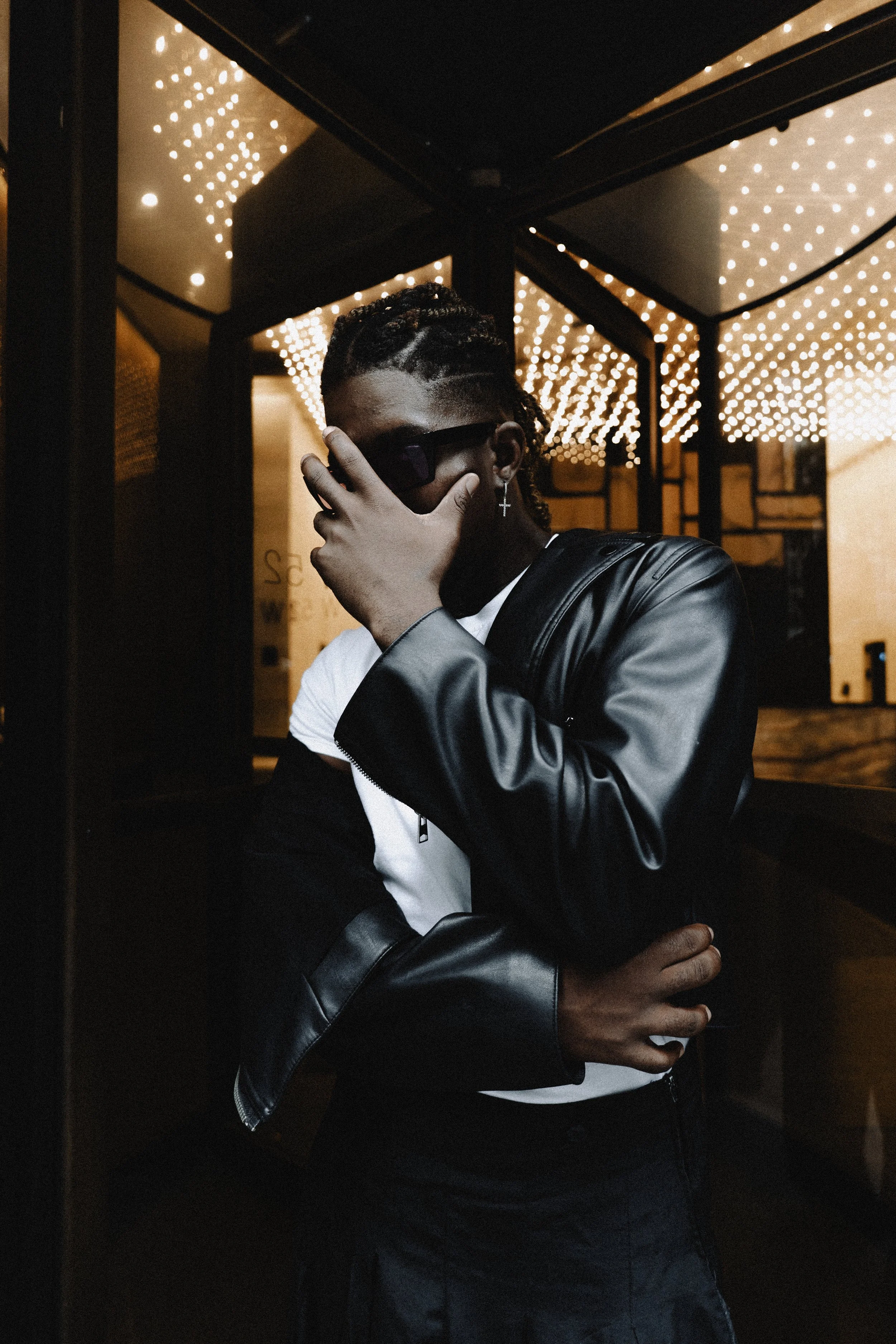 A person standing in an elevator with a black jacket, white shirt, and sunglasses, covering part of their face with their hand. The background features warm, ceiling lights reflected in the elevator's mirror.