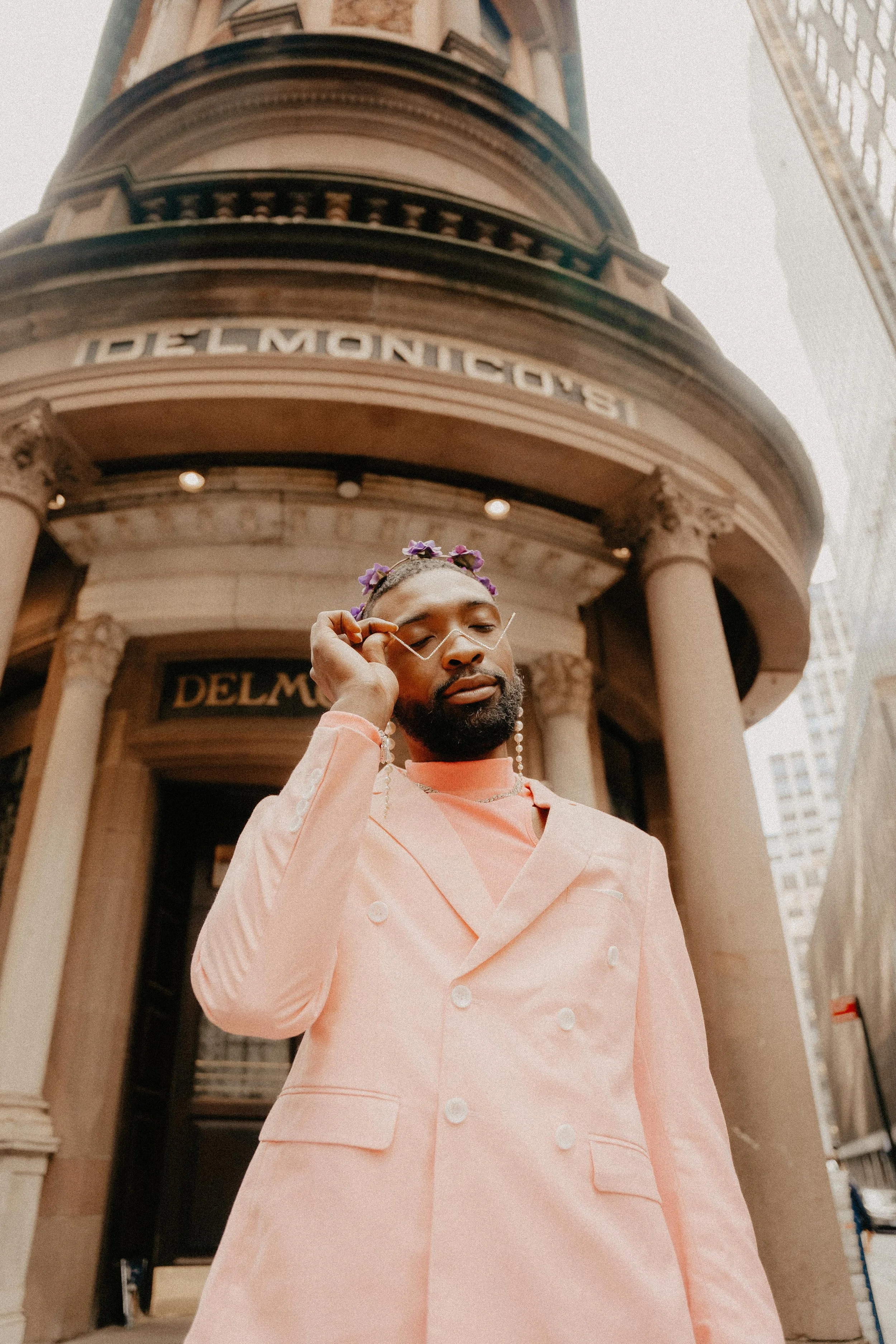 A man with a beard and floral headband adjusting glasses, standing in front of a building with the sign "Delmonico's" above the entrance, wearing a peach-colored chef's coat.