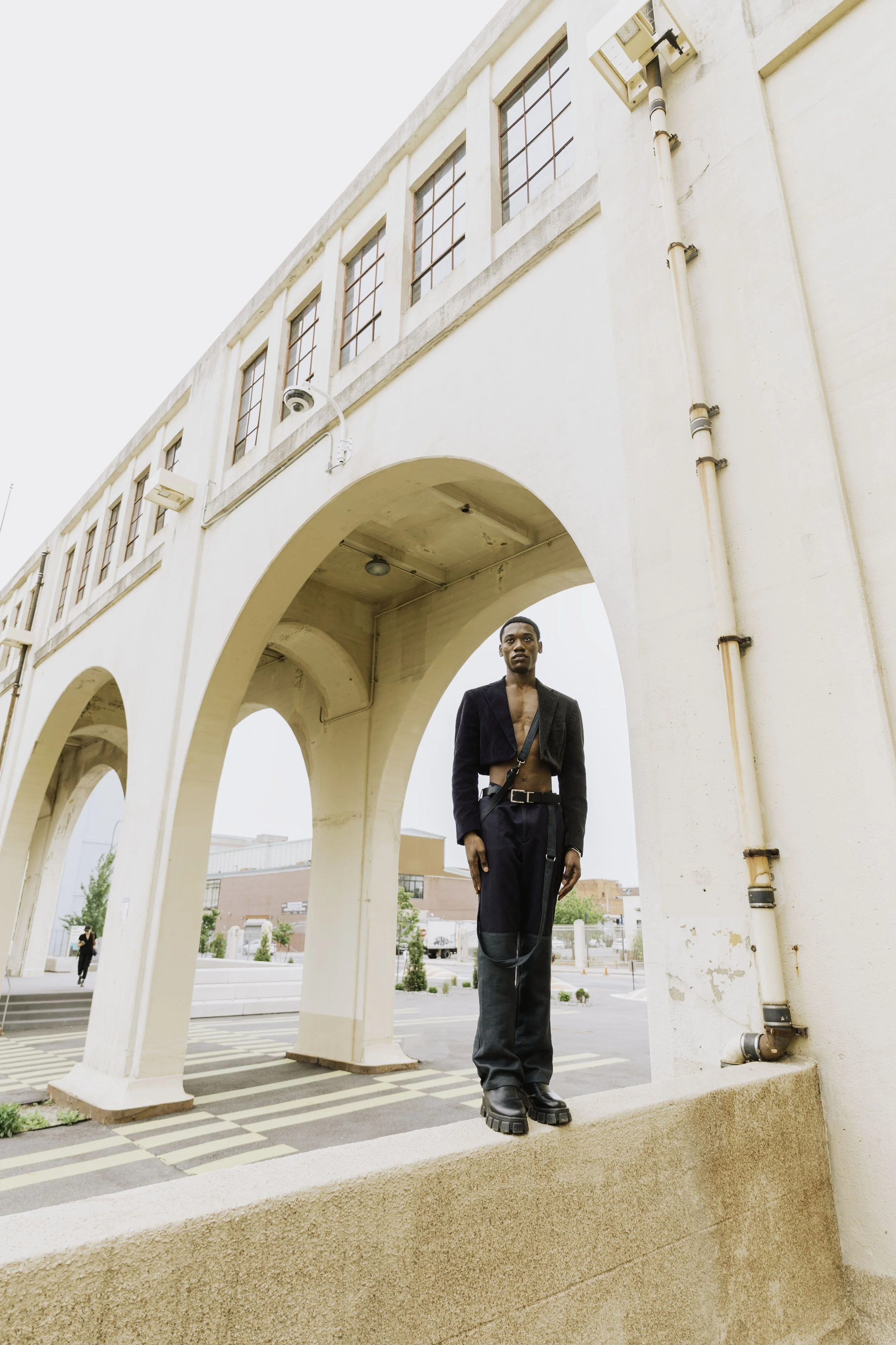 A person standing under a white archway of a building, wearing a black jacket and baggy pants, with a city street and buildings in the background.