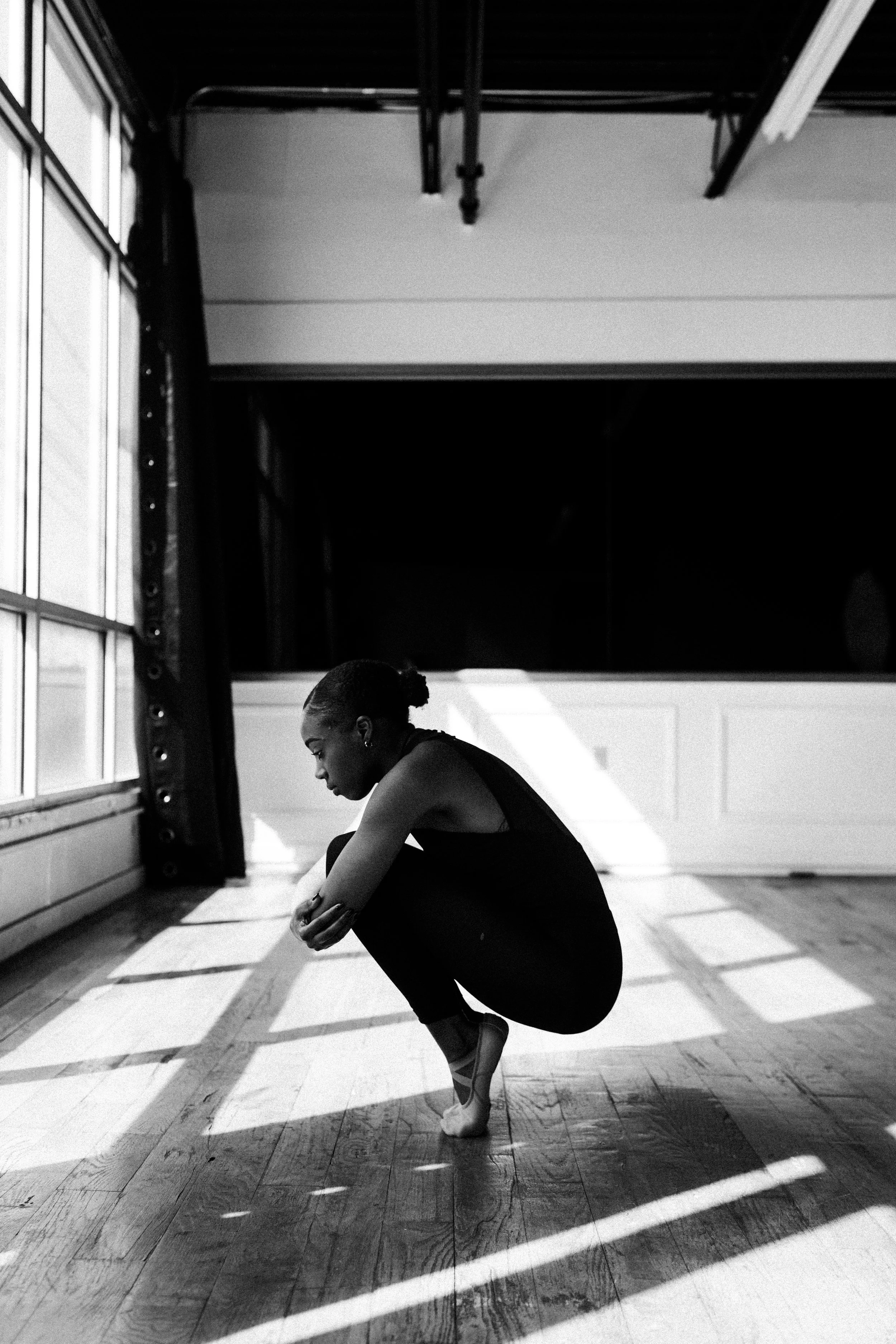 A woman in a gymnasium is practicing yoga, performing a pose where she is balancing on one foot while holding the toes of her other foot, with her head bowed and arms around her shin.
