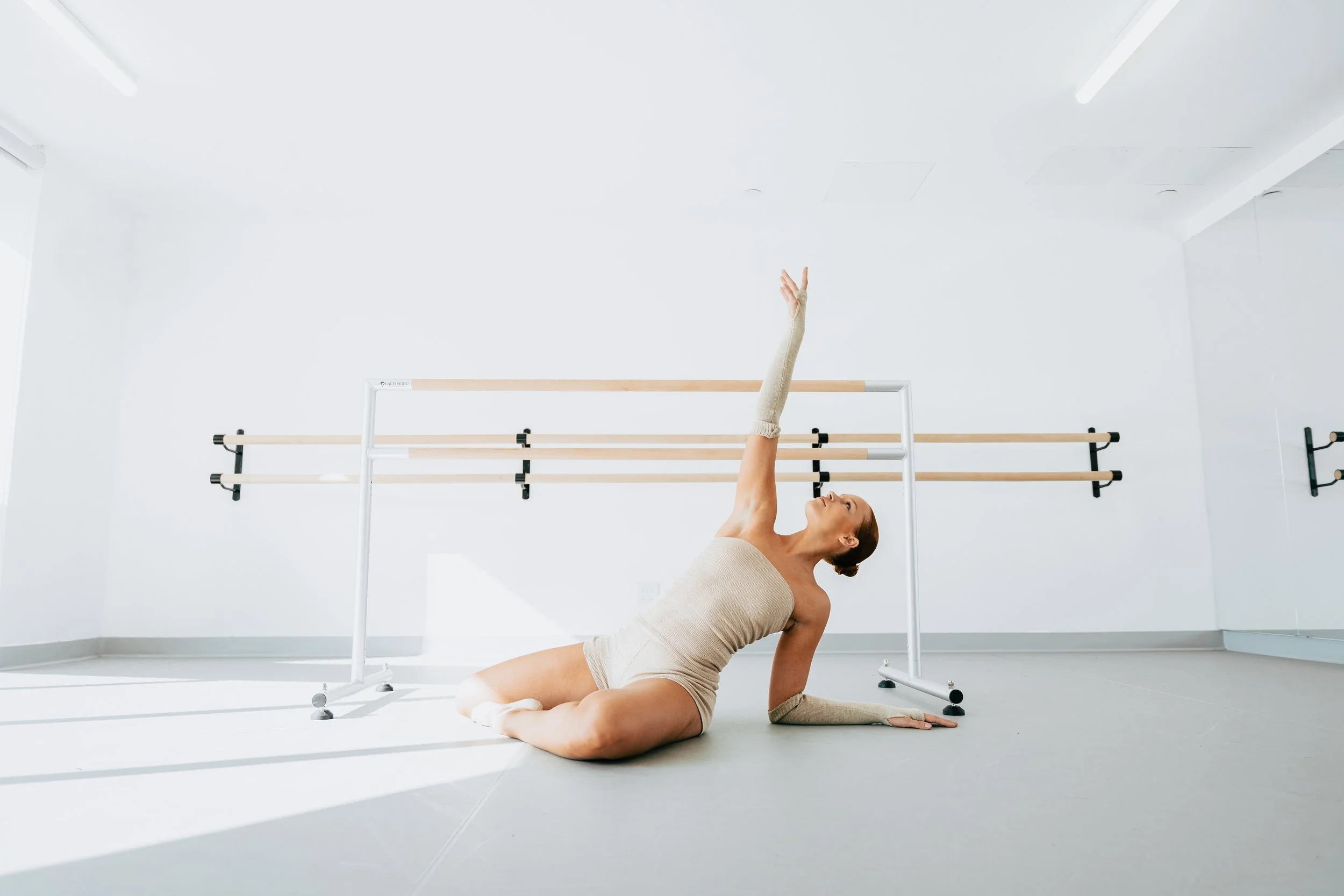 A woman in a beige workout outfit stretching on the floor in a bright, minimalistic exercise studio with ballet bars on the wall.