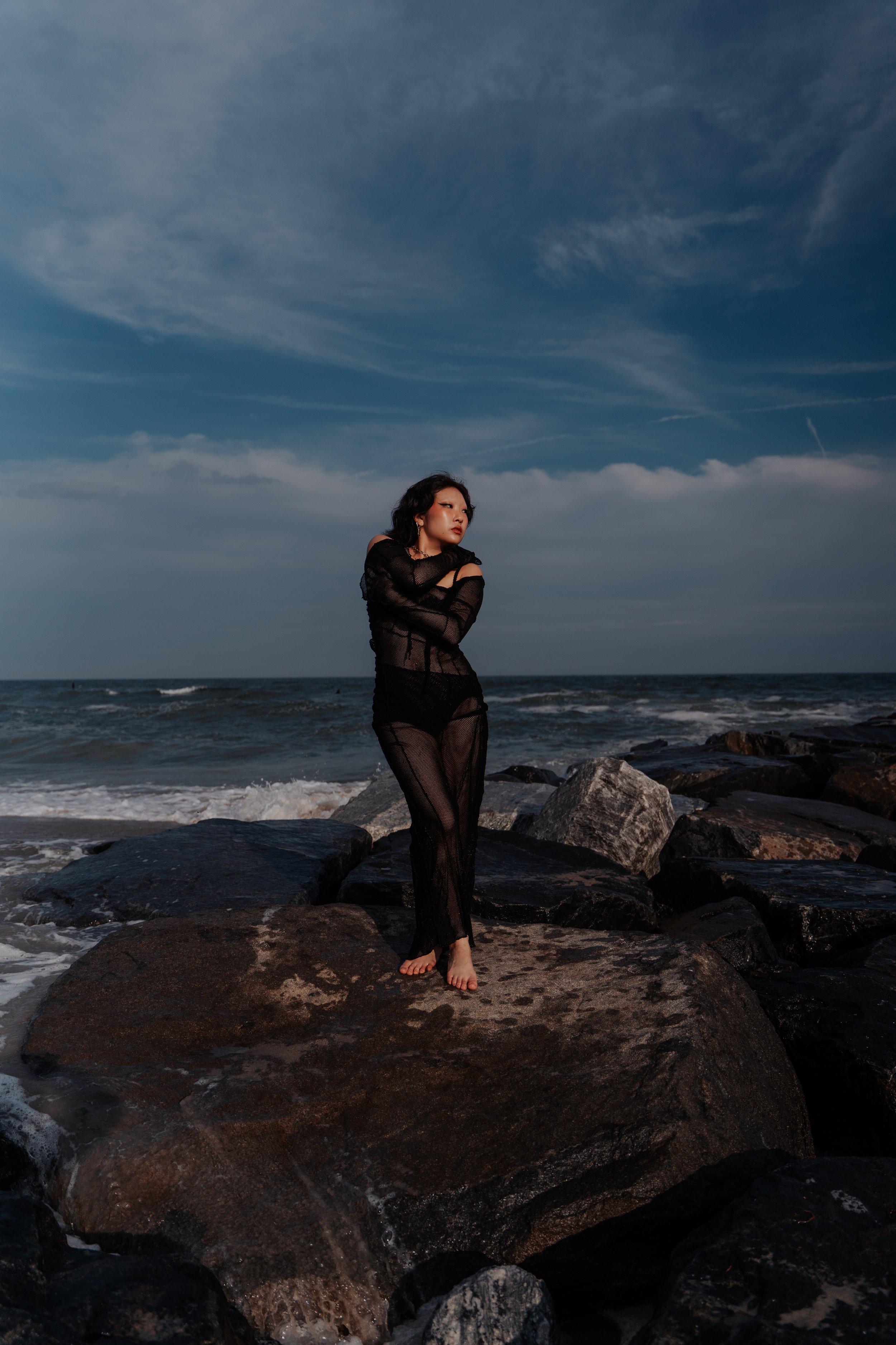 A woman standing on rocks at the beach during sunset or sunrise, wearing a sheer black dress, with the ocean and sky in the background.