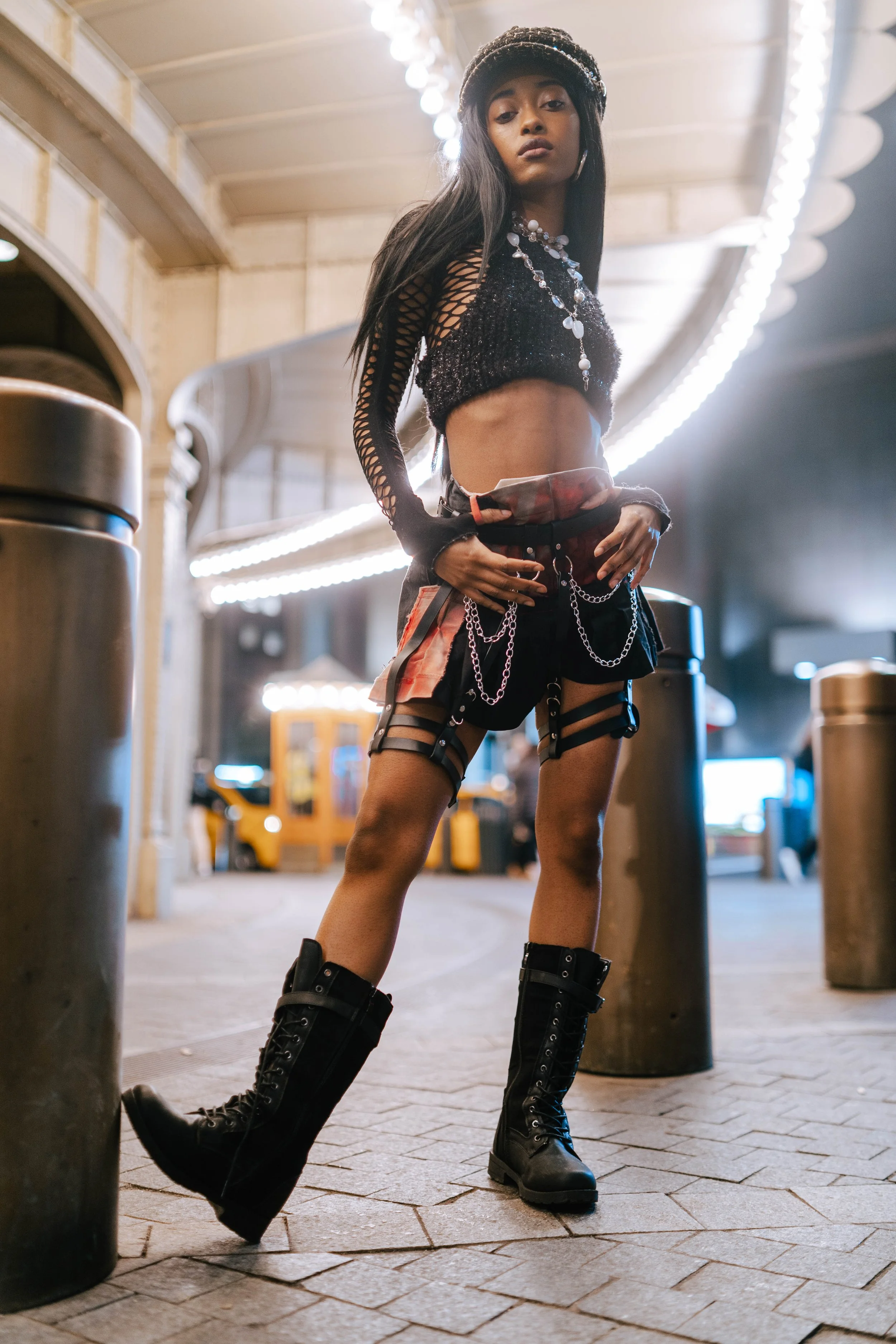 A young woman stands confidently in an urban setting, wearing a black mesh crop top with long sleeves, black shorts with chains, high lace-up boots, a beret, and jewelry, with bright lights above and concrete barriers around.