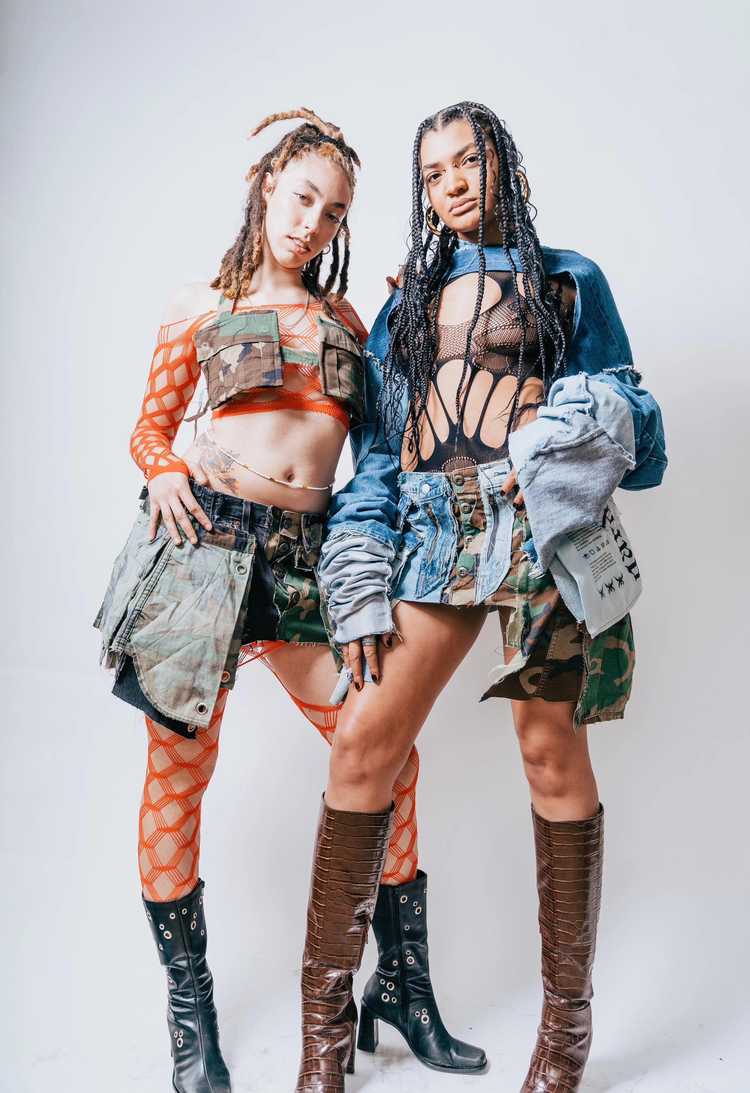 Two women standing together against a plain white background, wearing edgy, eclectic fashion outfits with camouflage, denim, and fishnet details, and high boots.