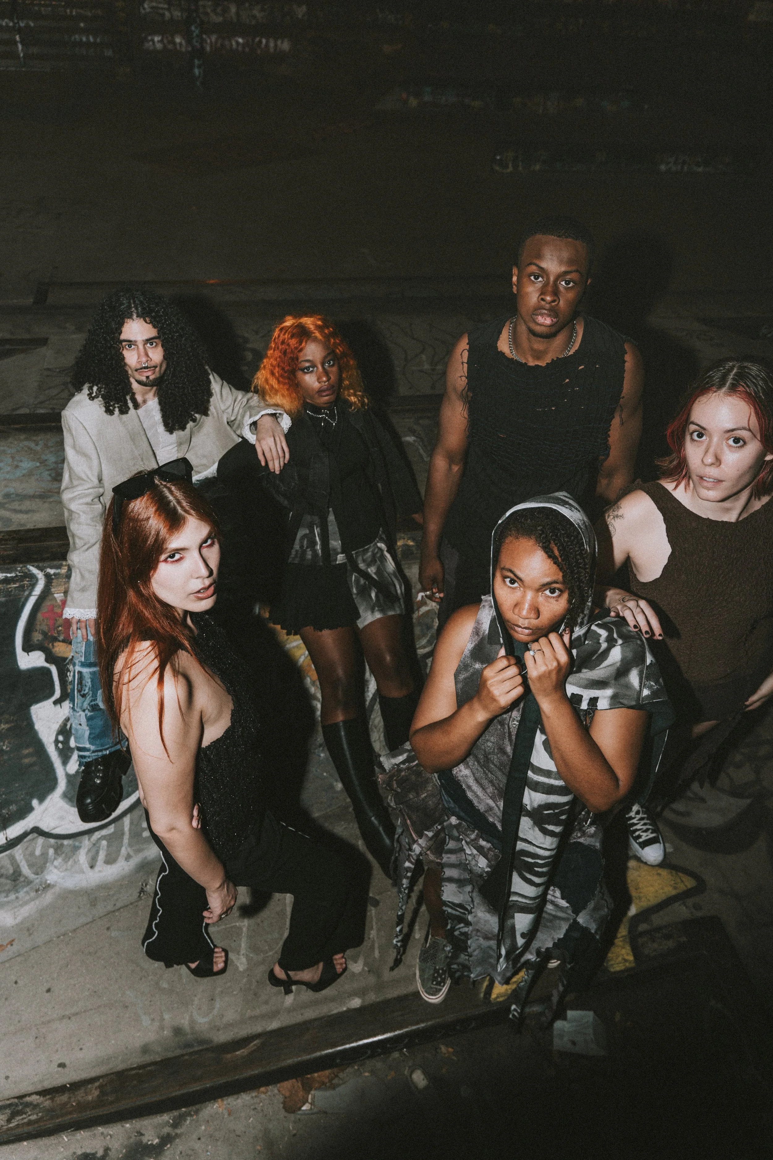 A group of six diverse young people posing together in an urban setting. Some are standing, some are sitting, and they are dressed in fashionable, edgy clothing with bold hairstyles and makeup. The background features graffiti and dark scenery.