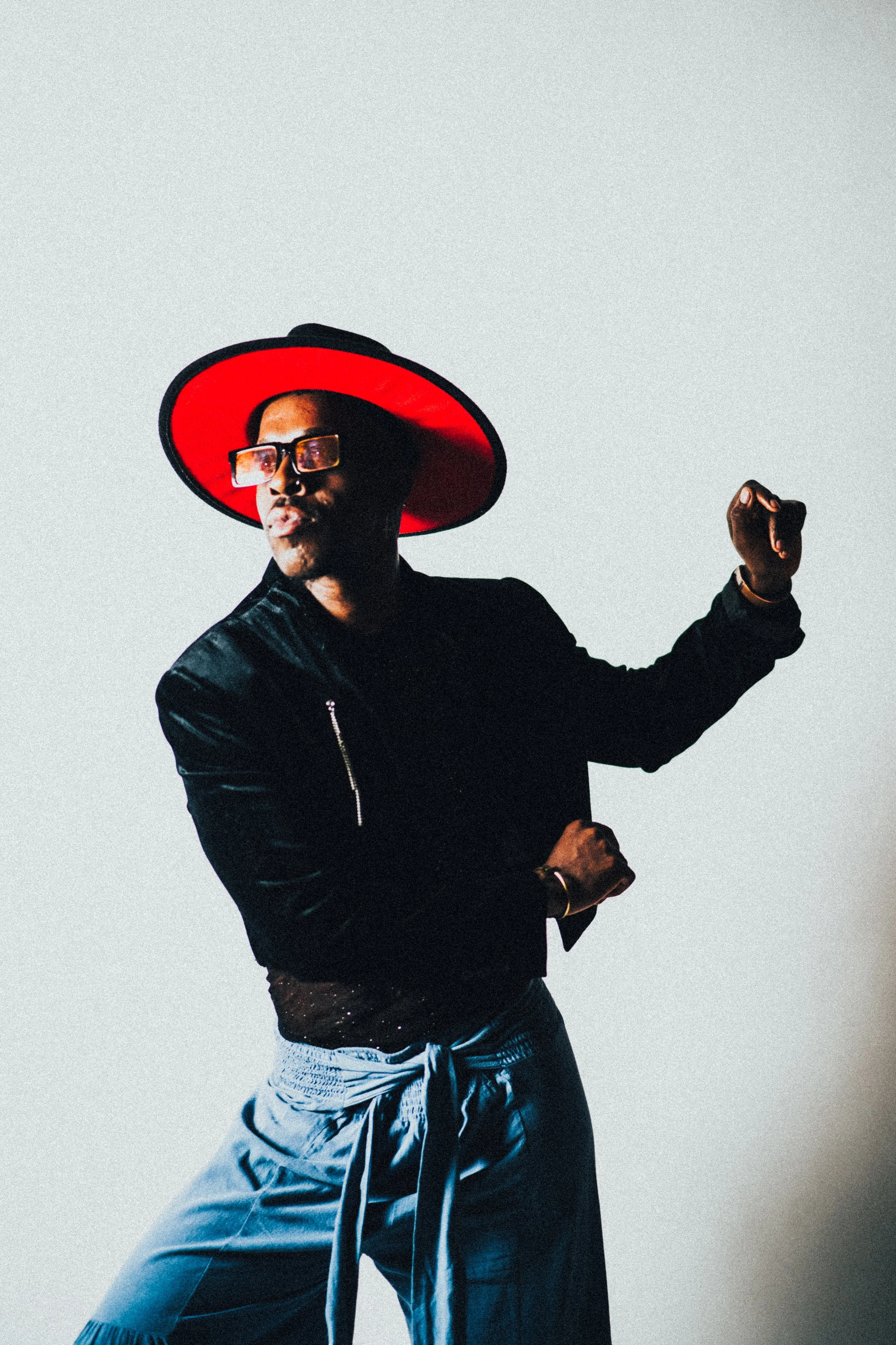 A person wearing a large red and black wide-brimmed hat, sunglasses, a black leather jacket, and patterned pants, standing against a plain light background with a confident pose and raised arm.