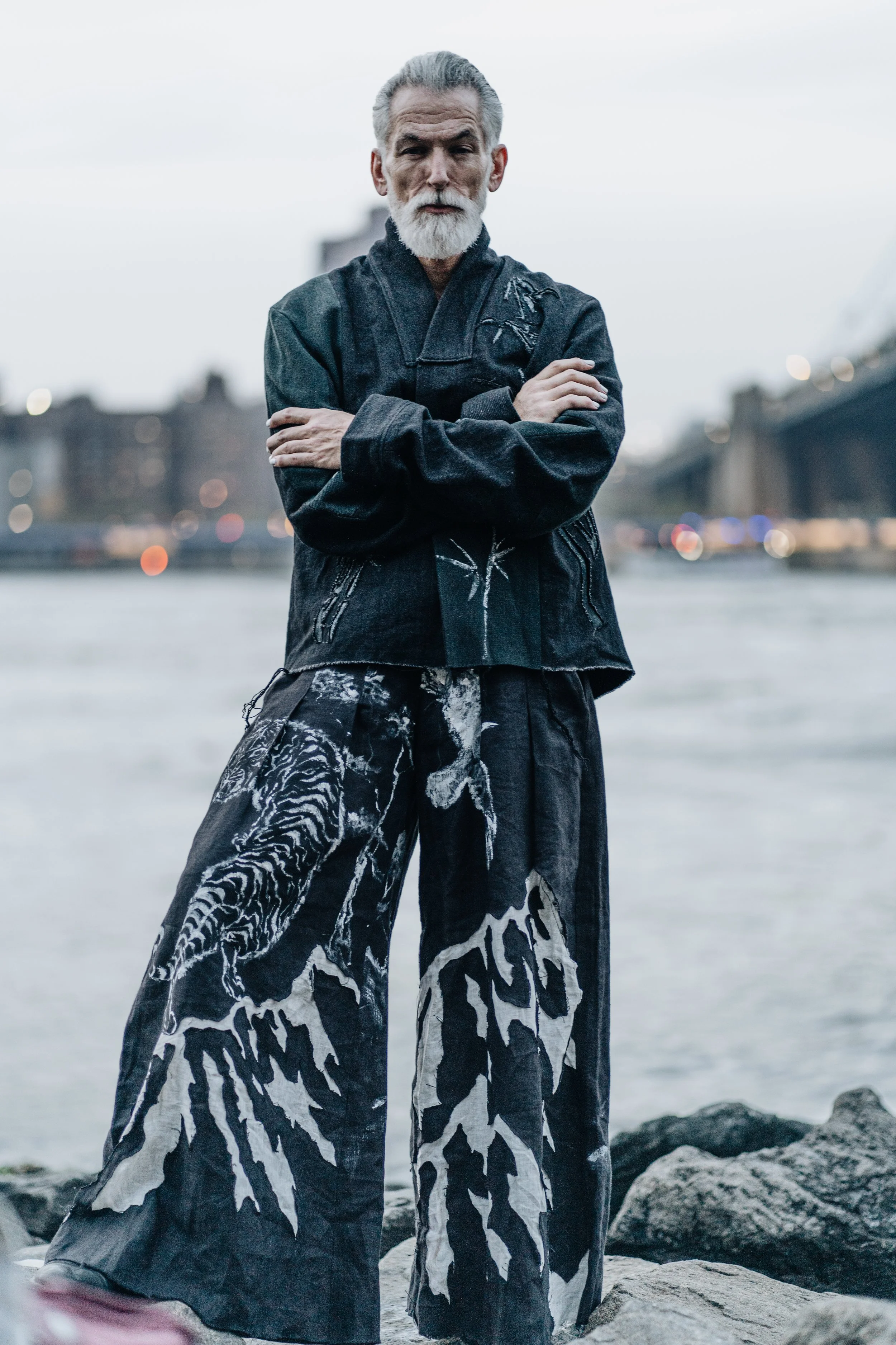 An older man with gray hair and beard wearing a black jacket with artistic embroidery, standing outdoors near a body of water with rocks, with an overcast sky and blurred cityscape in the background.