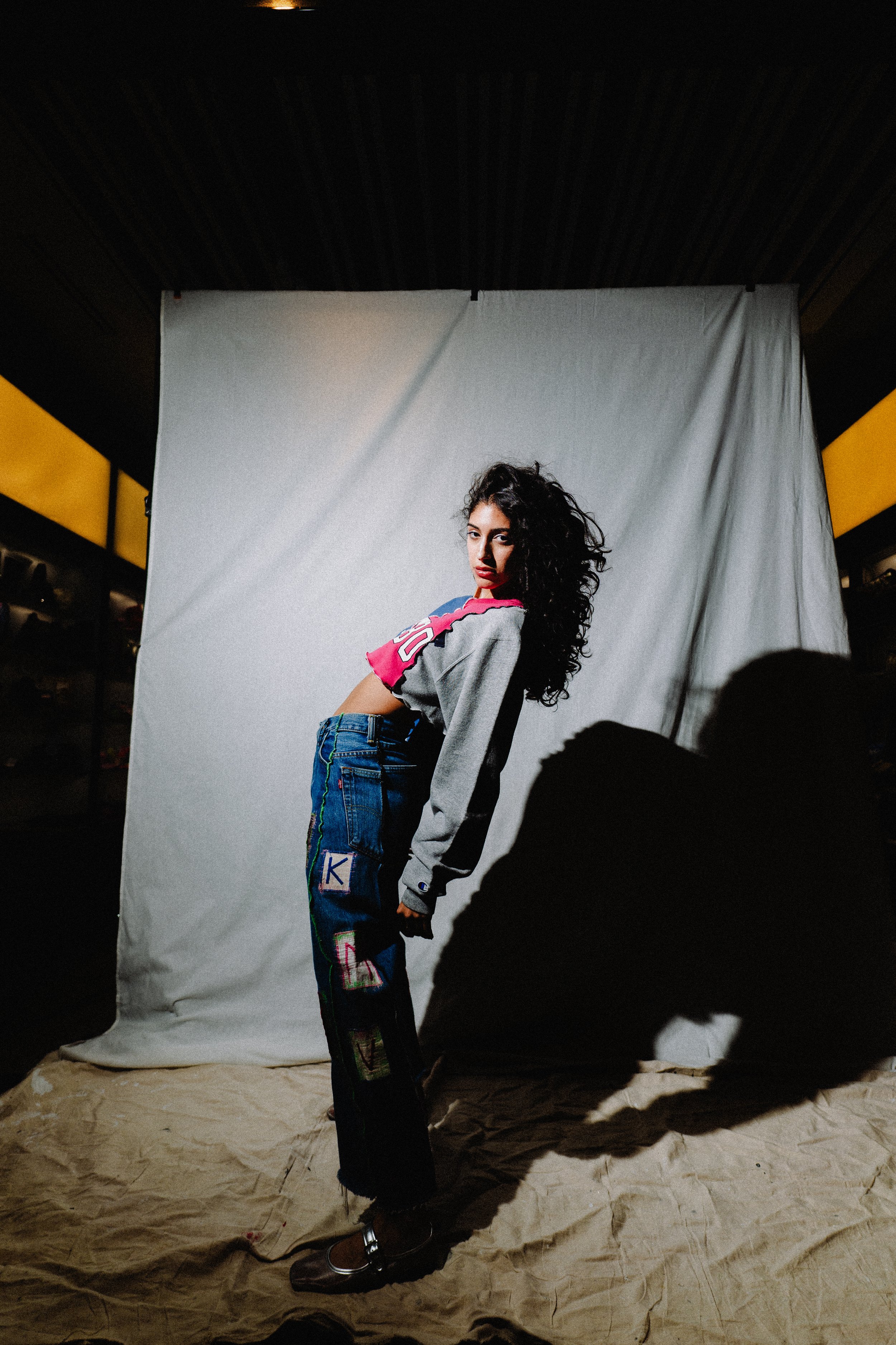 A young woman with long, curly dark hair poses in front of a plain white backdrop. She is wearing a gray jacket with pink and purple accents, blue patchwork jeans with colorful patches, and brown shoes. Shadows cast on crumpled beige paper on the flo