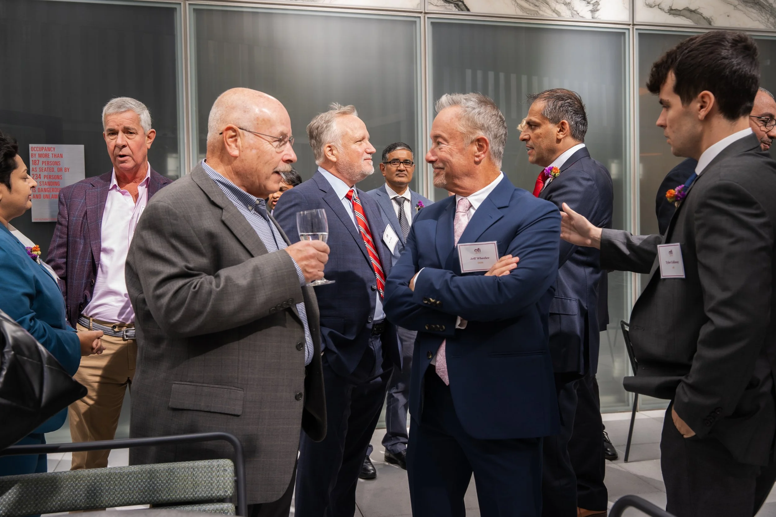 Group of professionally dressed men and women engaged in conversation at an indoor event, some wearing name tags and small flower boutonnieres.
