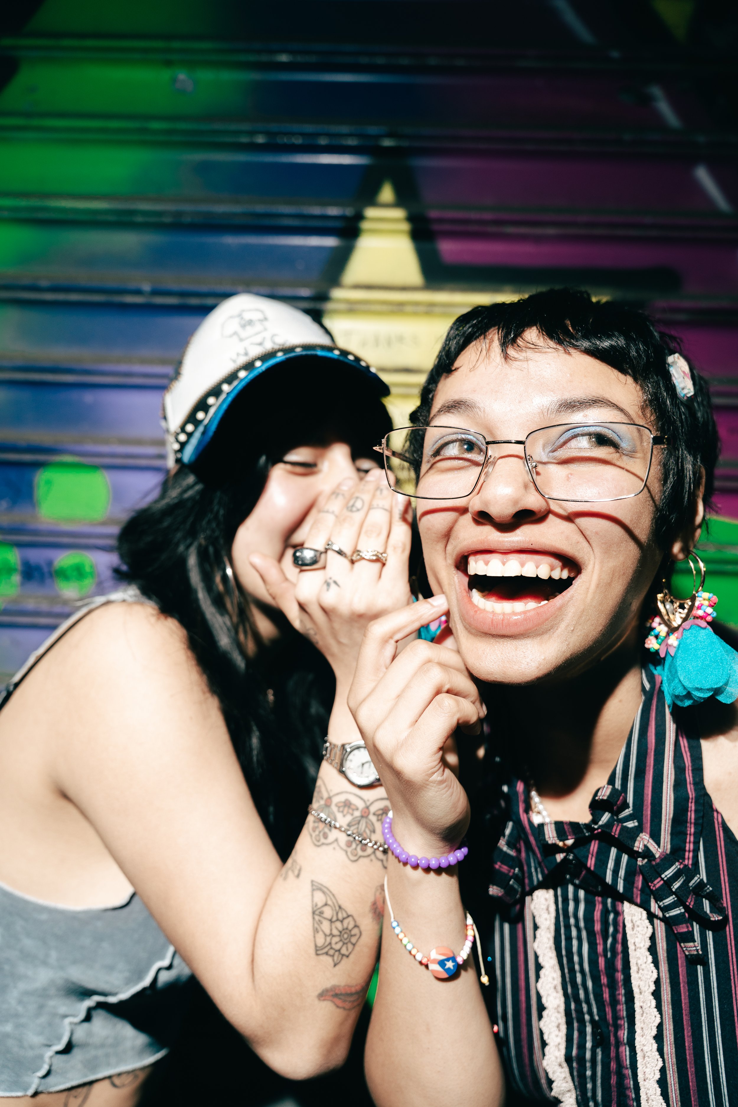 Two people smiling and laughing closely together, with one whispering into the other's ear against a colorful, graffiti-covered background.