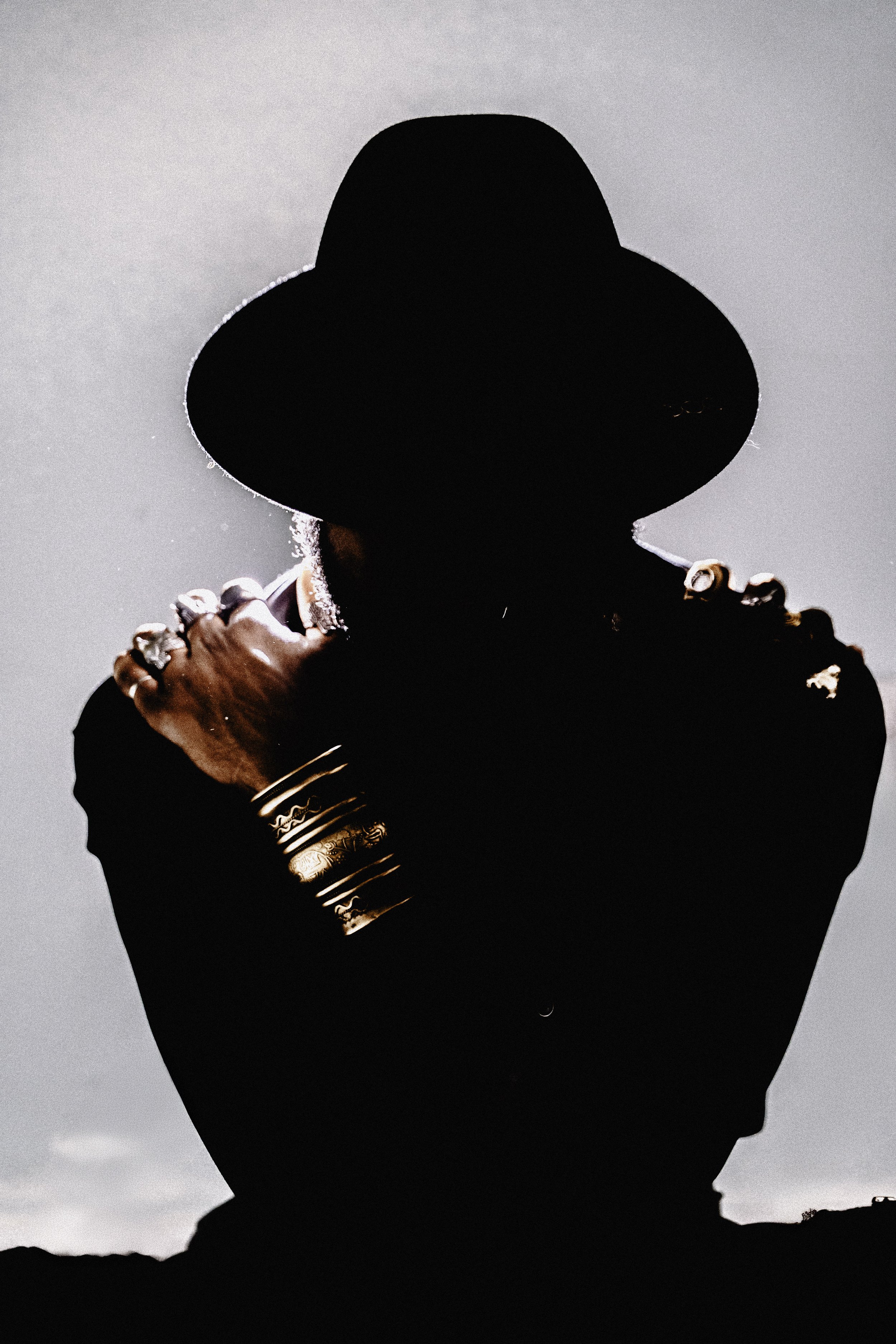 Silhouette of a person wearing a wide-brimmed hat, adorned with rings and bangles, against a cloudy sky.