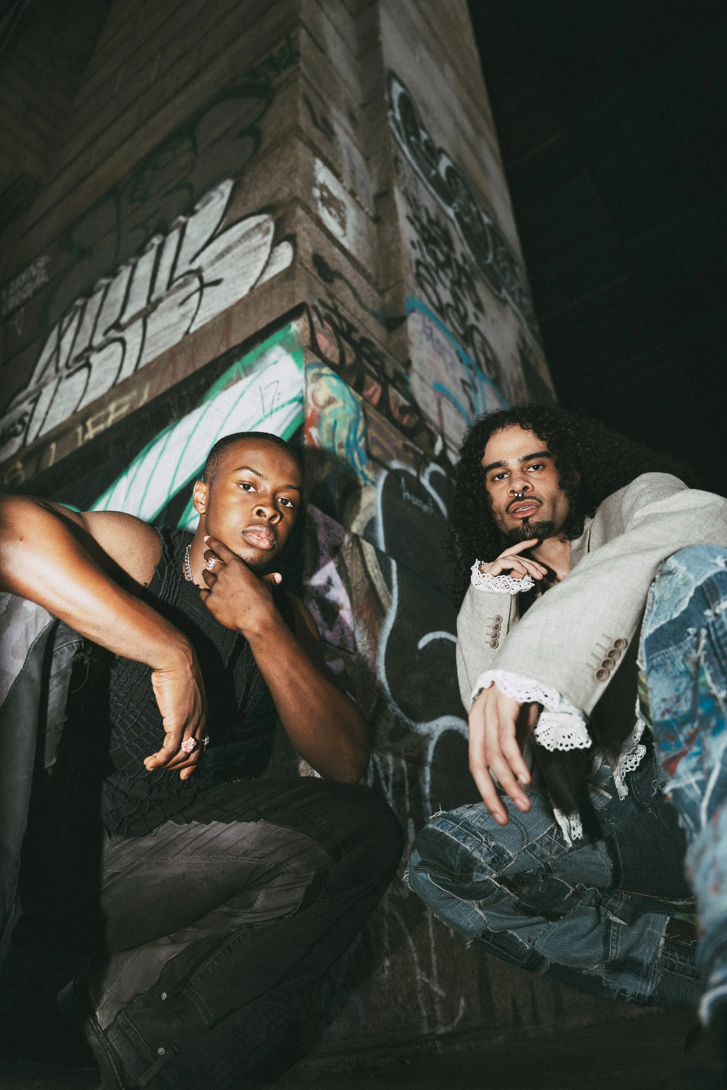 Two young adults, a woman with braided hair and a man with curly hair, sitting close together against a graffiti-covered brick wall at night.