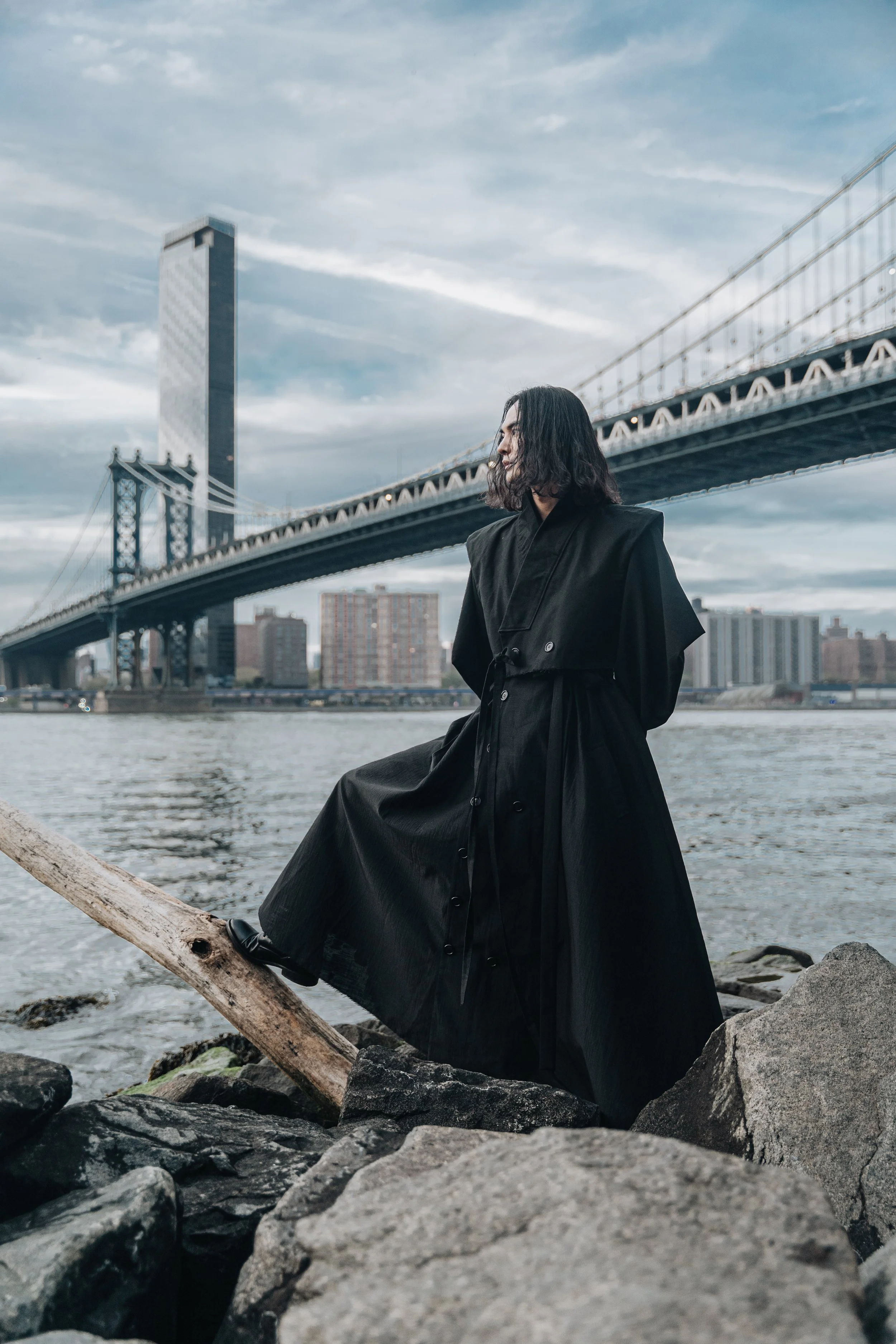 A woman with shoulder-length black hair wearing a black coat sitting on rocks by the river with a city skyline and a bridge in the background on a cloudy day.